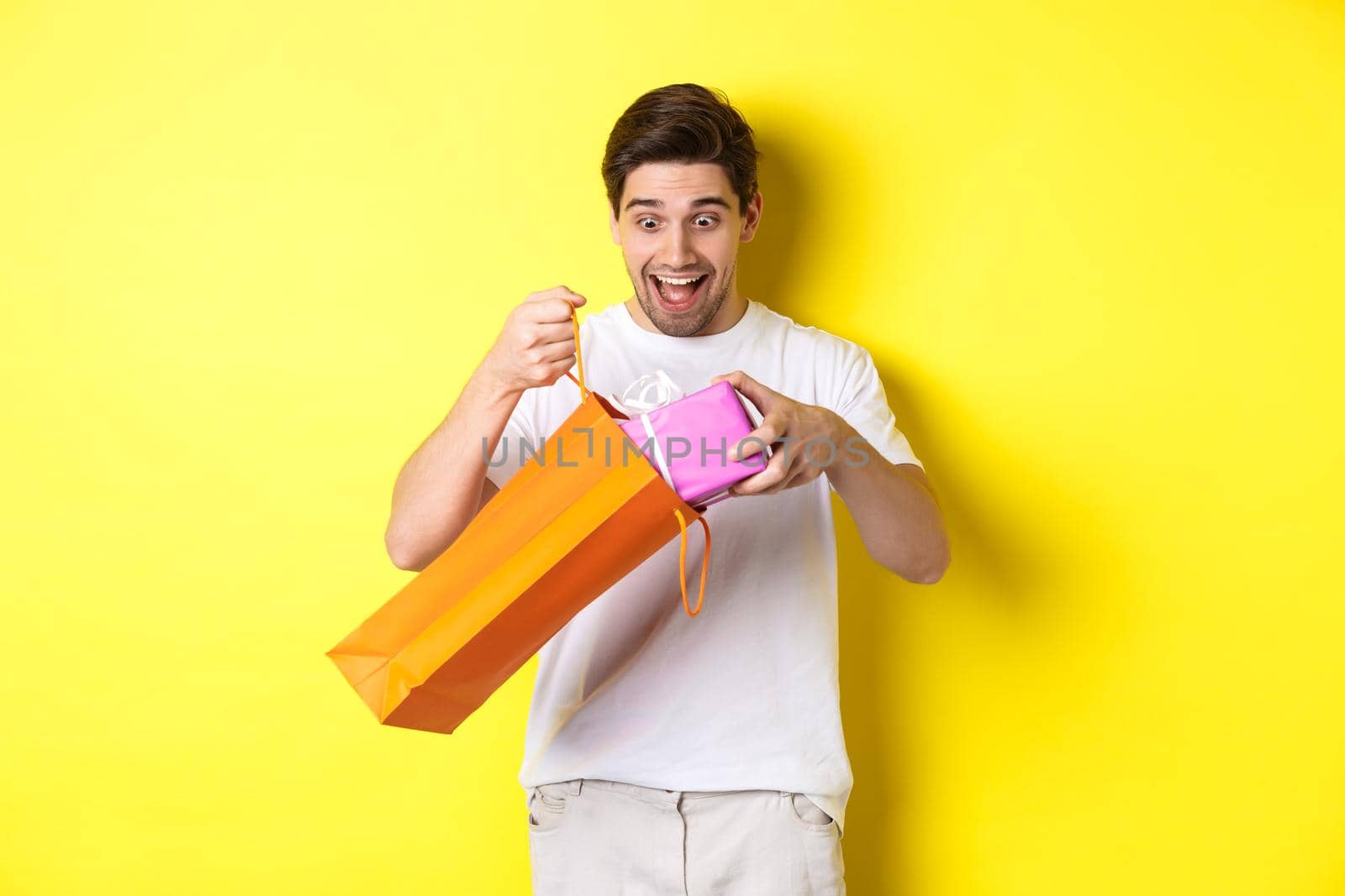 Concept of holidays and celebration. Young man looking surprised as take out gift from shopping bag, standing over yellow background.