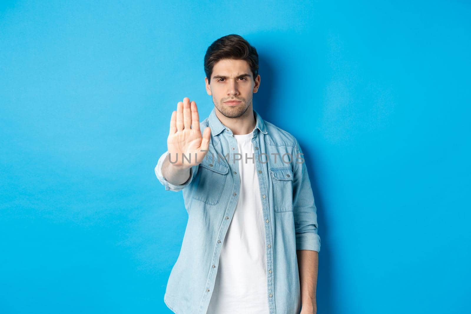 Serious man frowning and saying no, extending hand to shop stop sign, prohibit action, standing against blue background by Benzoix