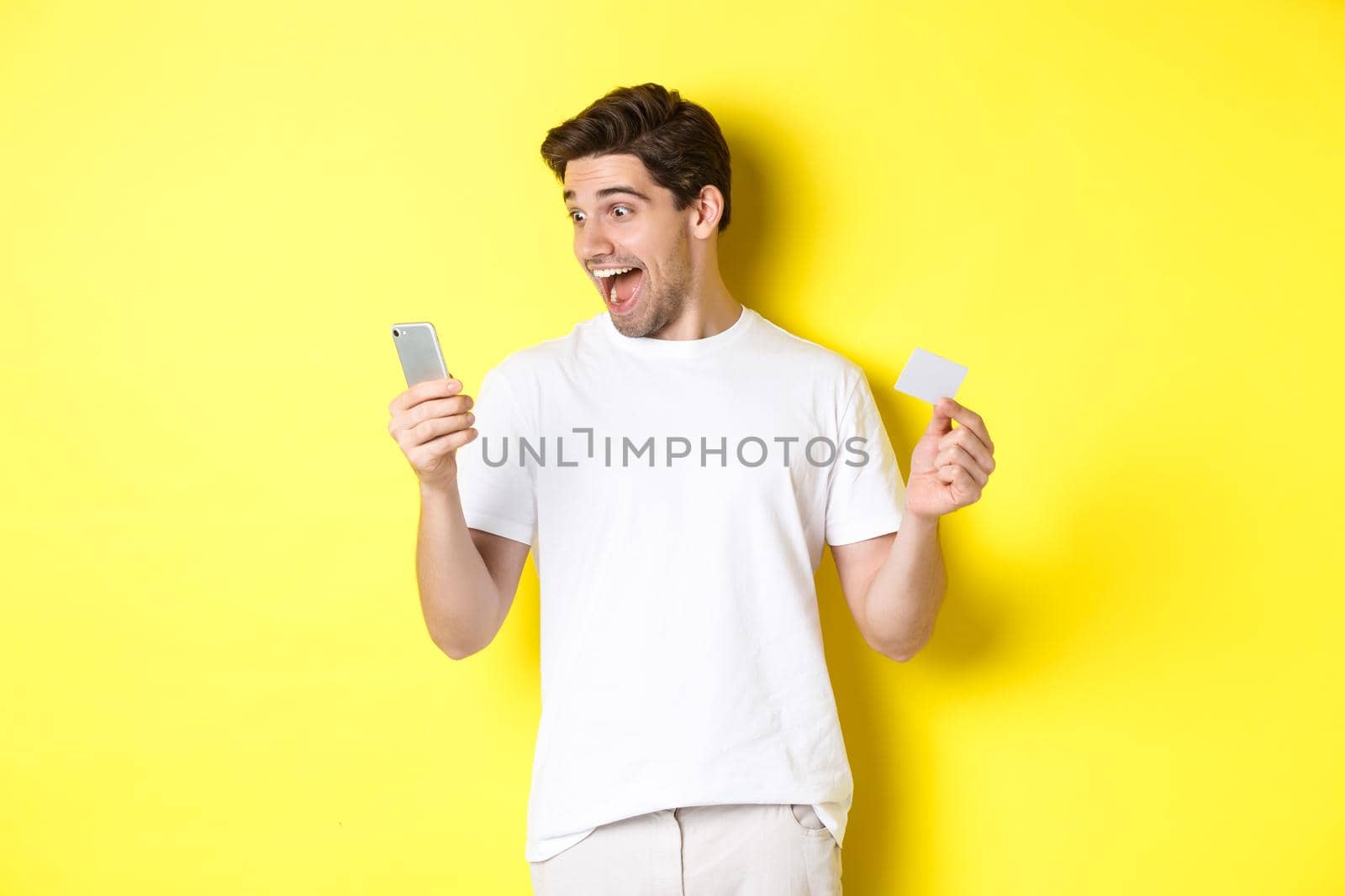 Surprised guy holding smartphone and credit card, online shopping on black friday, standing over yellow background by Benzoix