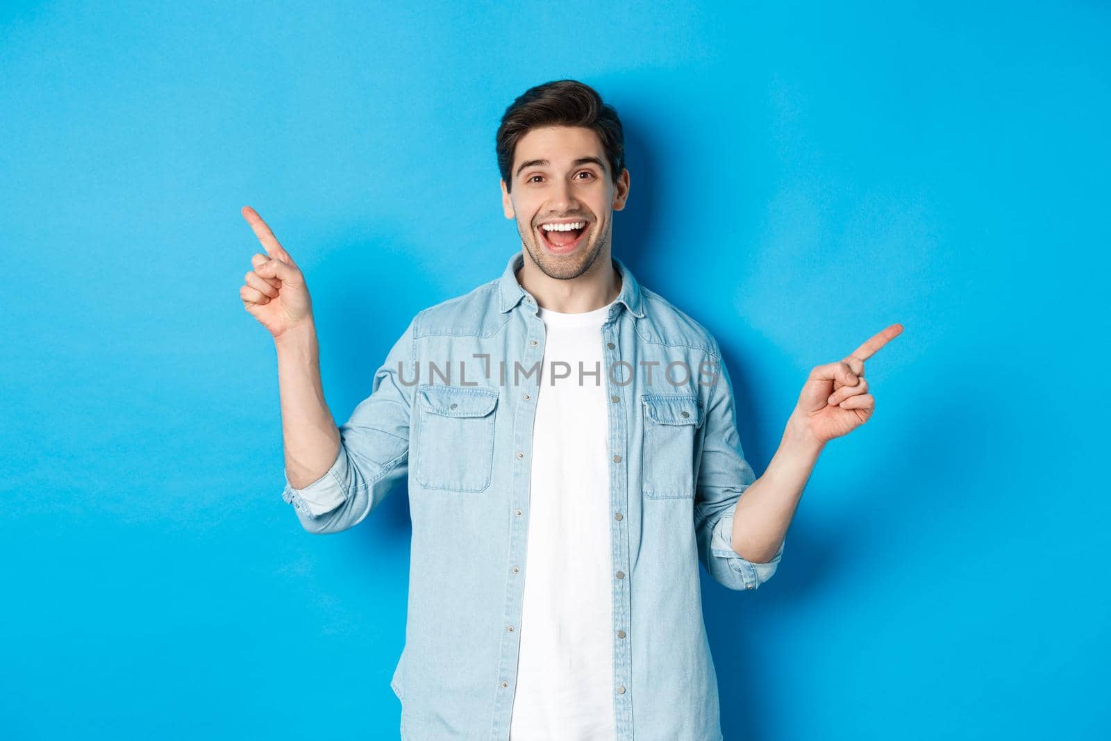 Cheerful adult man smiling, pointing fingers sideways, showing left and right promo banners, standing against blue background.