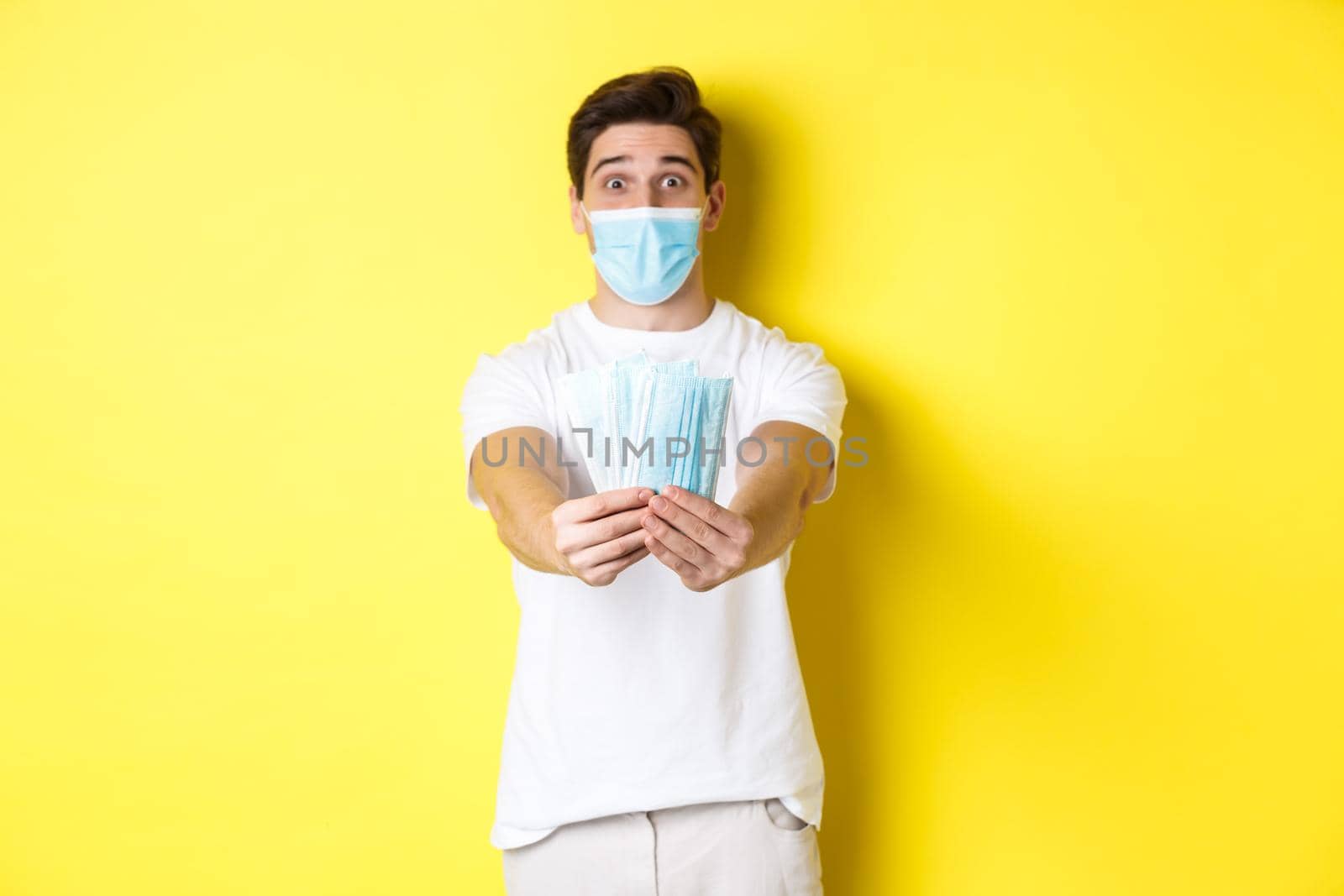 Concept of covid-19, quarantine and preventive measures. Young caucasian man giving medical masks for you, standing against yellow background.