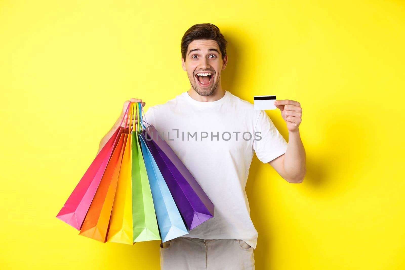 Happy attractive man holding shopping bags, showing credit card, standing over yellow background by Benzoix