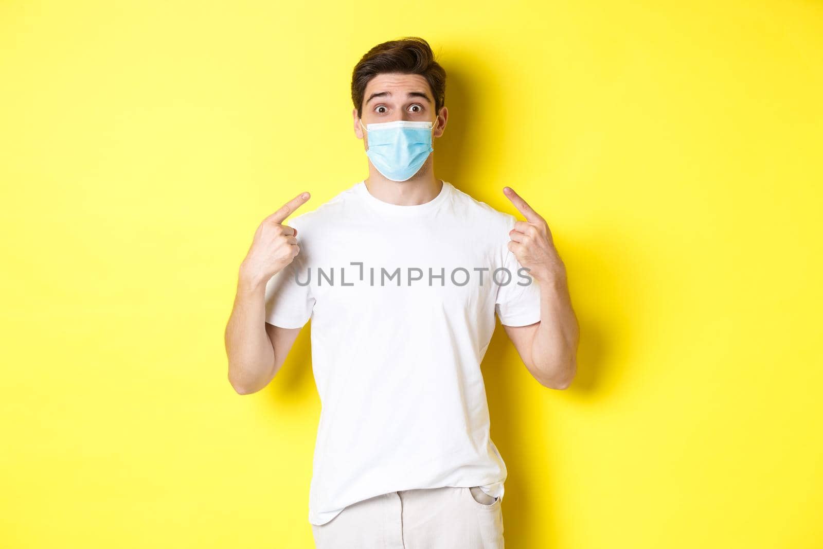 Concept of coronavirus, pandemic and social distancing. Young surprised man pointing at medical mask on face, yellow background by Benzoix