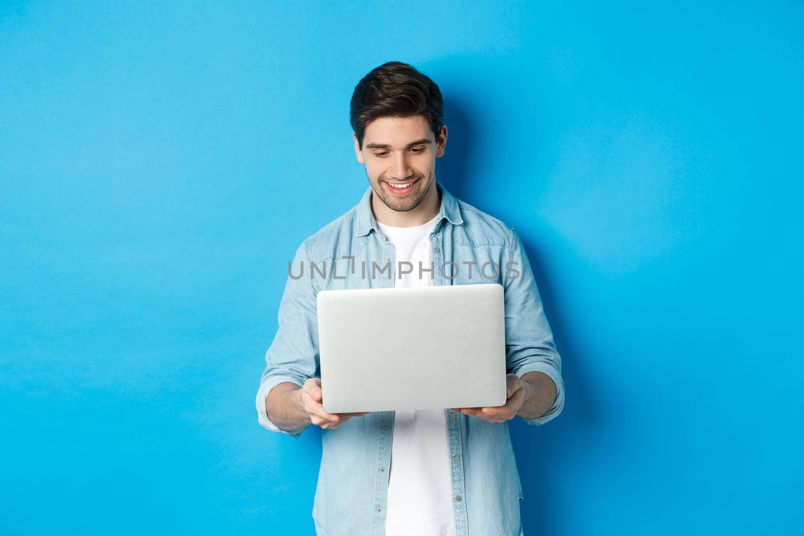 Handsome man working on laptop, smiling and looking at screen satisfied, standing against blue background.