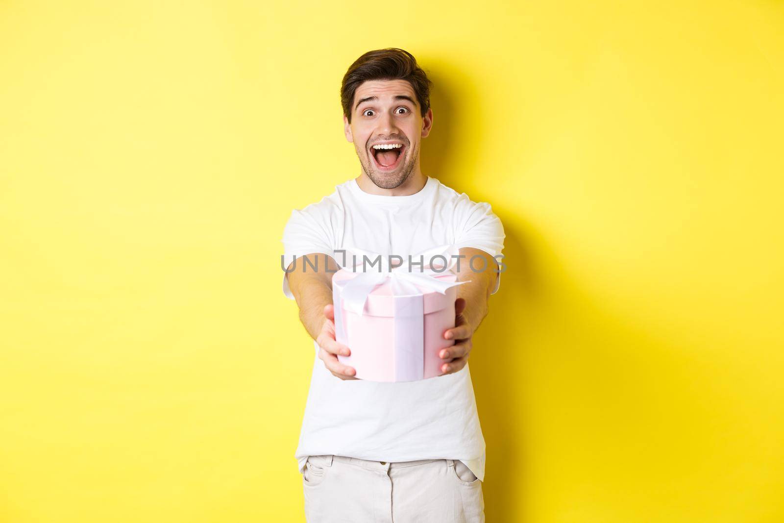 Concept of holidays and celebration. Happy man wishing happy new year, giving you gift, standing over yellow background by Benzoix