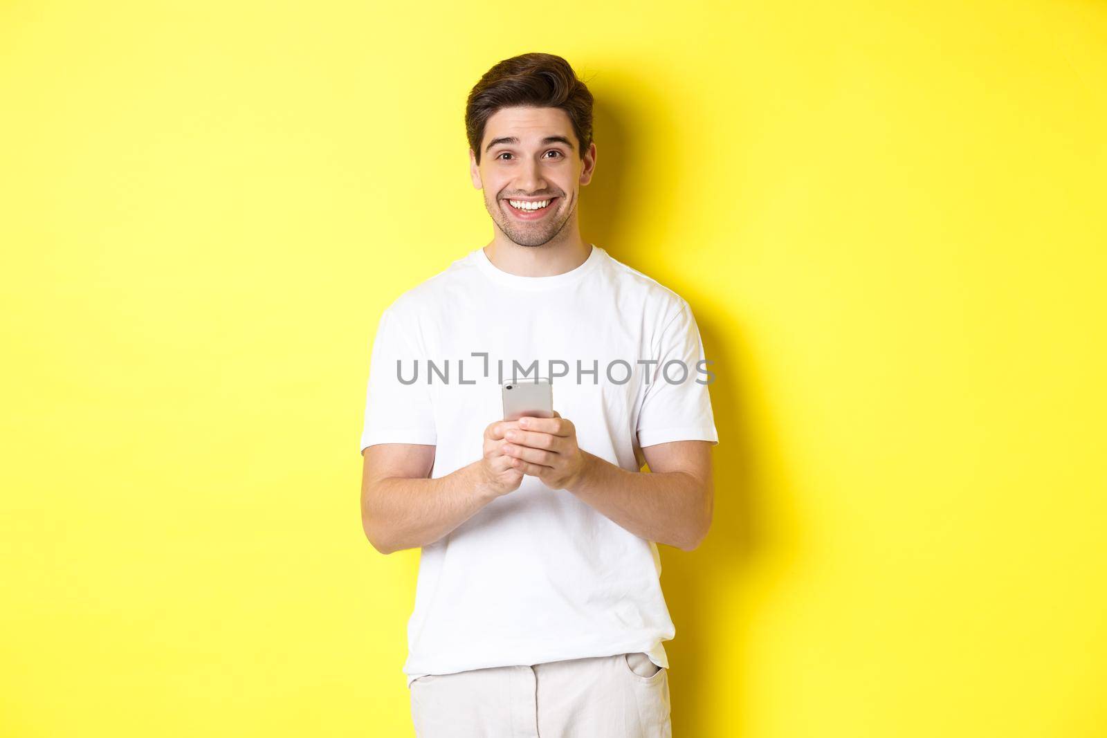 Man smiling and looking happy after reading promo offer on smartphone, standing against yellow background in white t-shirt by Benzoix