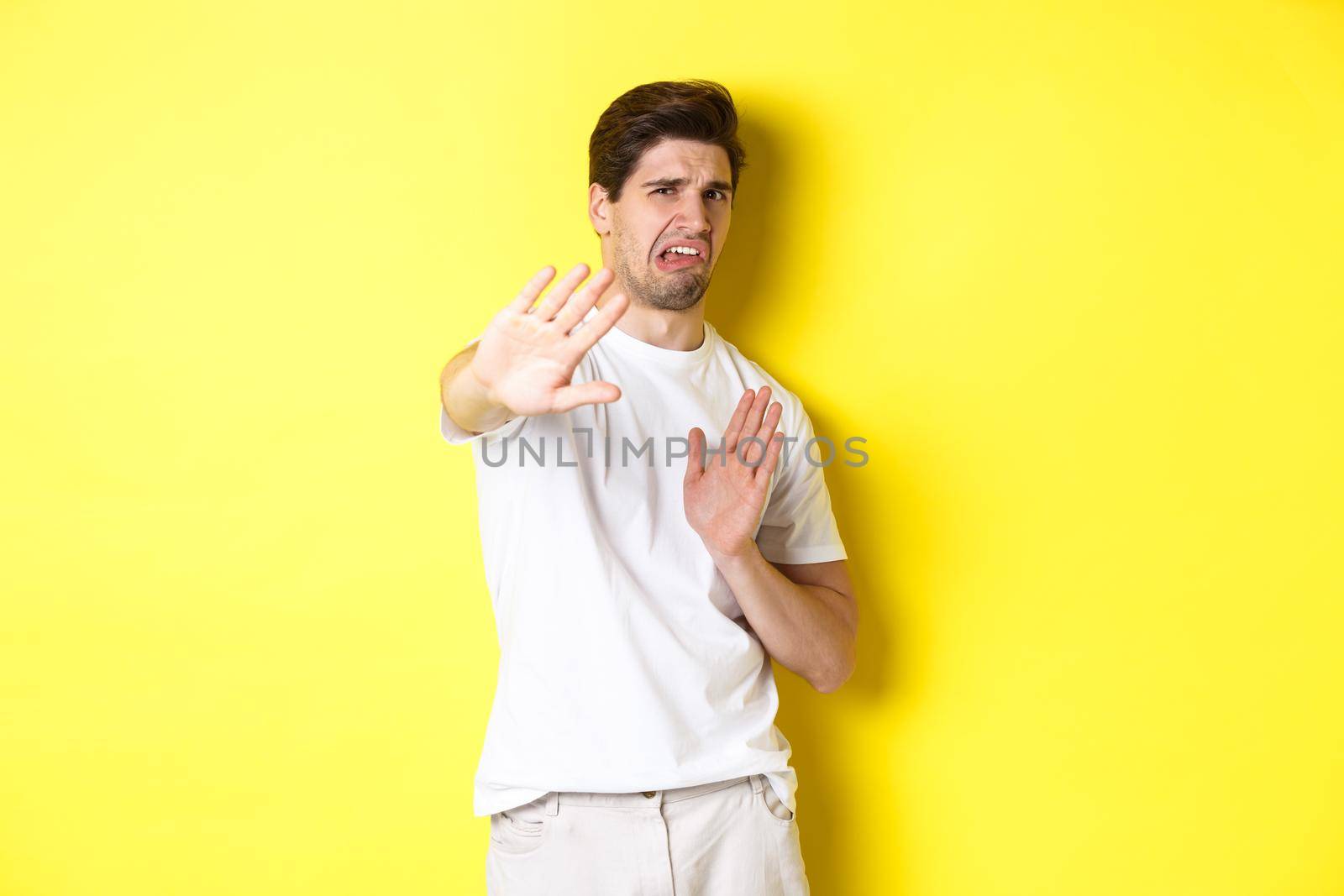 Disgusted man refusing, grimacing from dislike and aversion, begging to stop, standing in white t-shirt against yellow background.