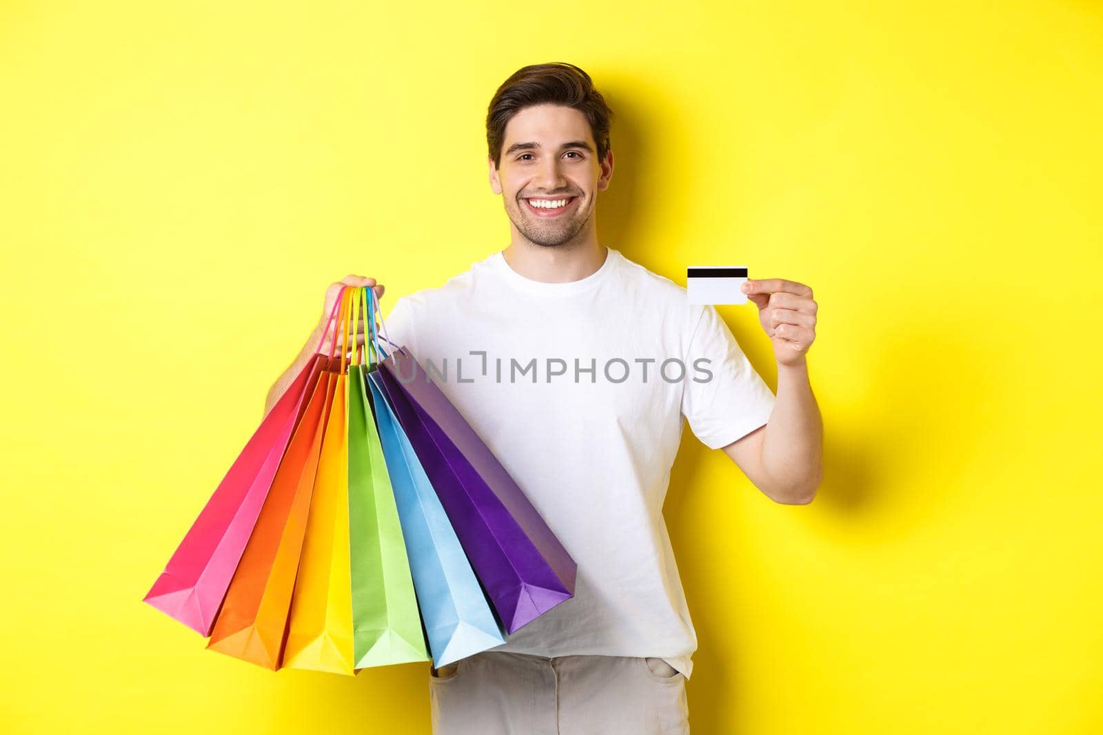 Young man shopping for holidays, holding paper bags and recommending bank credit card, standing over yellow background.