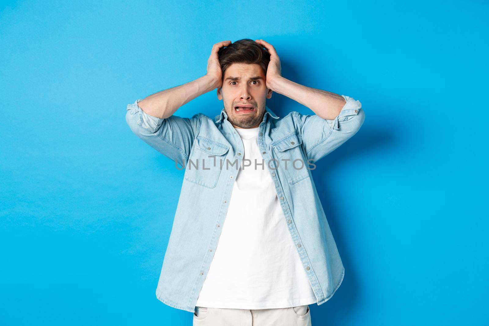 Image of man in panic holding hands on head, looking frustrated and anxious, standing against blue background.