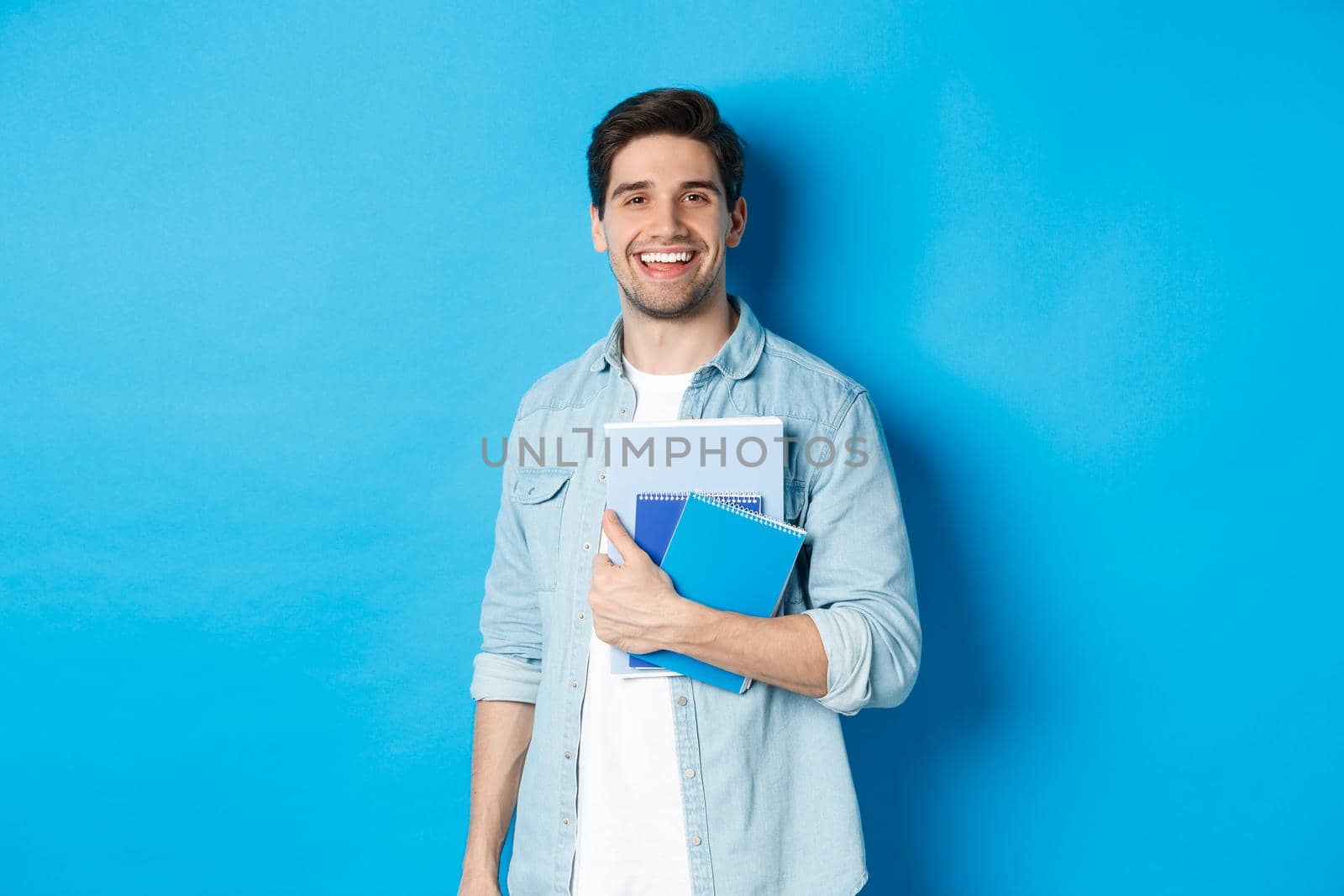 Smiling man studying, holding notebooks and looking happy, standing over blue background.
