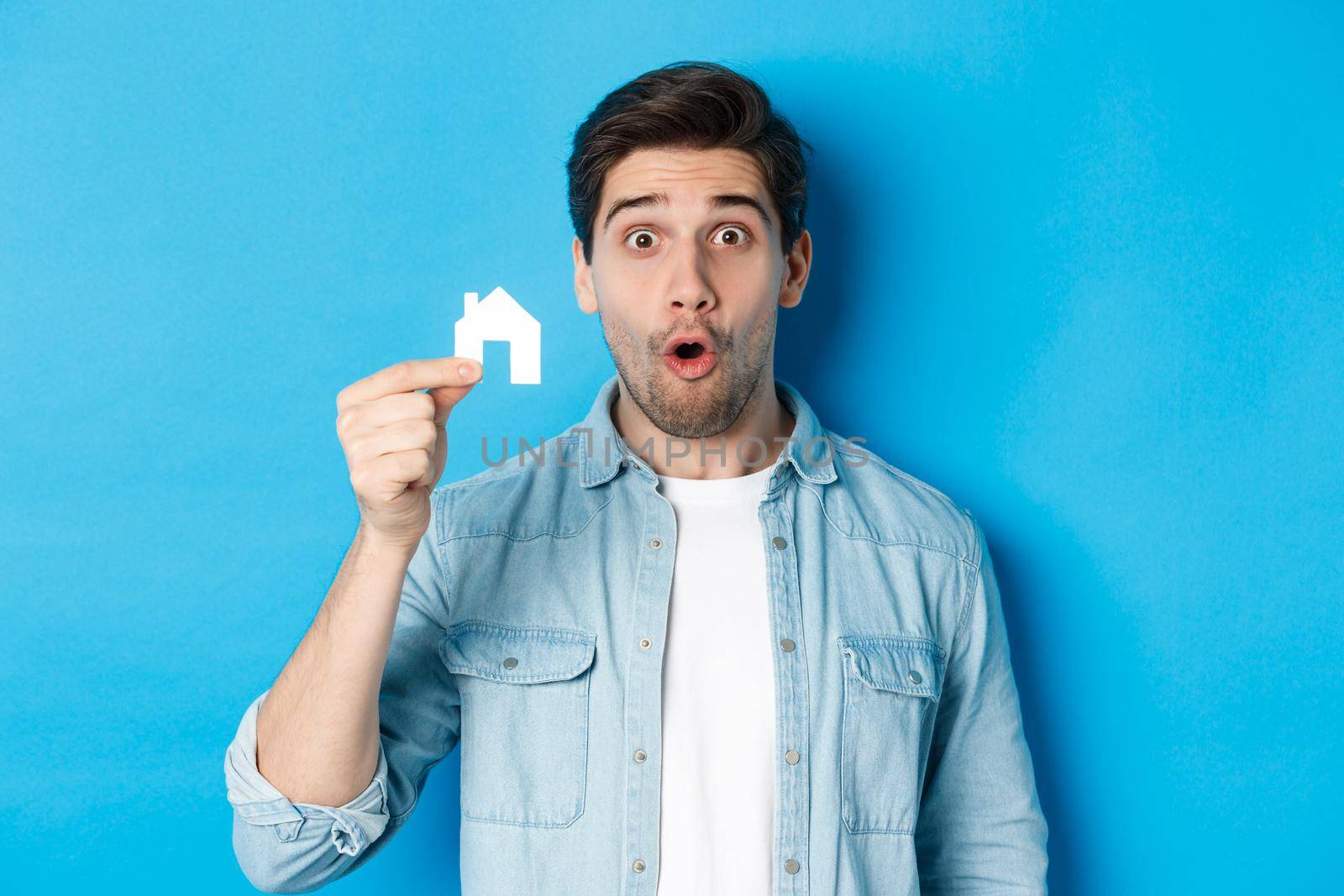 Real estate concept. Surprised guy showing small paper house model, looking amazed, standing against blue background by Benzoix