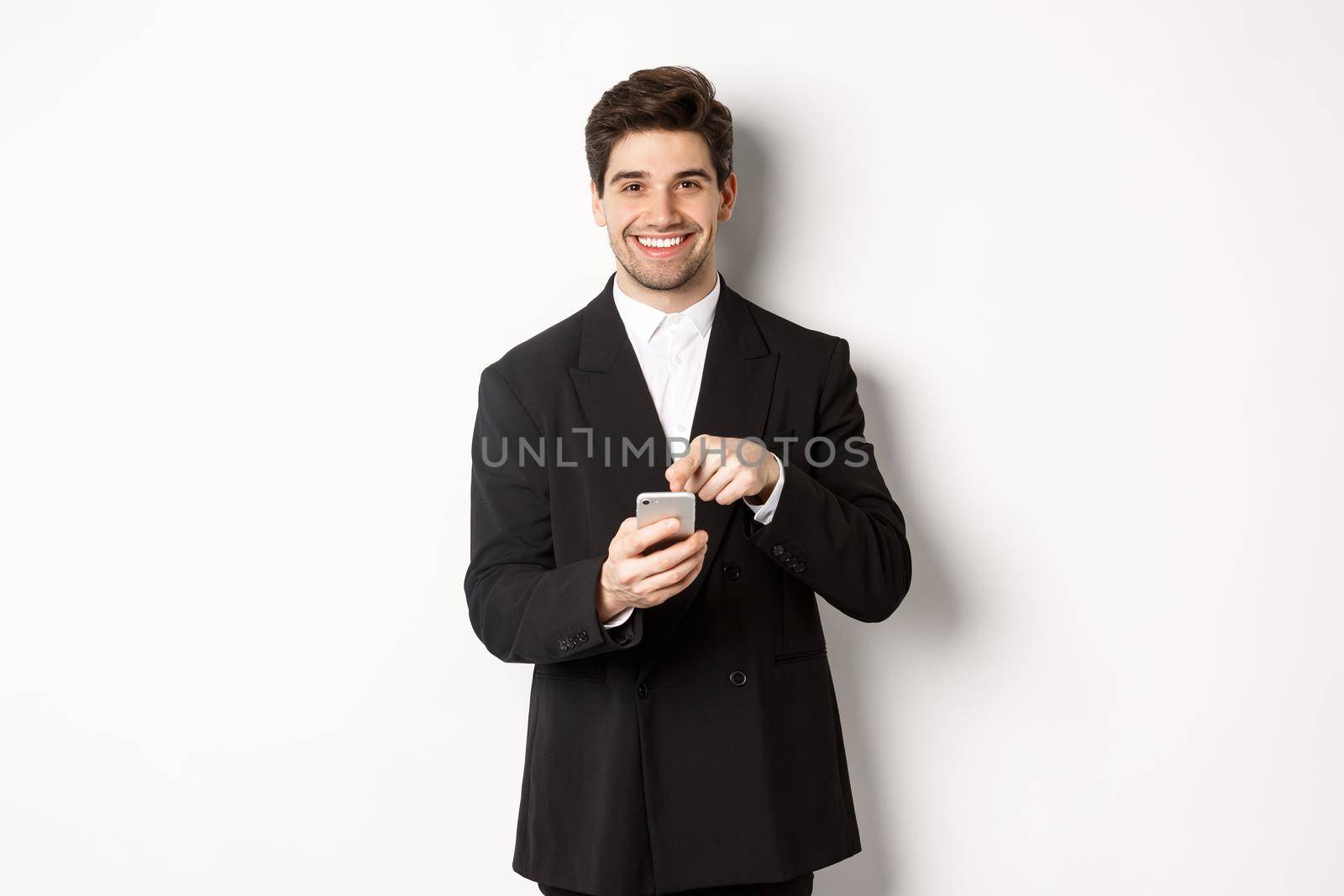 Image of handsome businessman in suit showing you app on smartphone, pointing finger at mobile screen and smiling, standing over white background by Benzoix