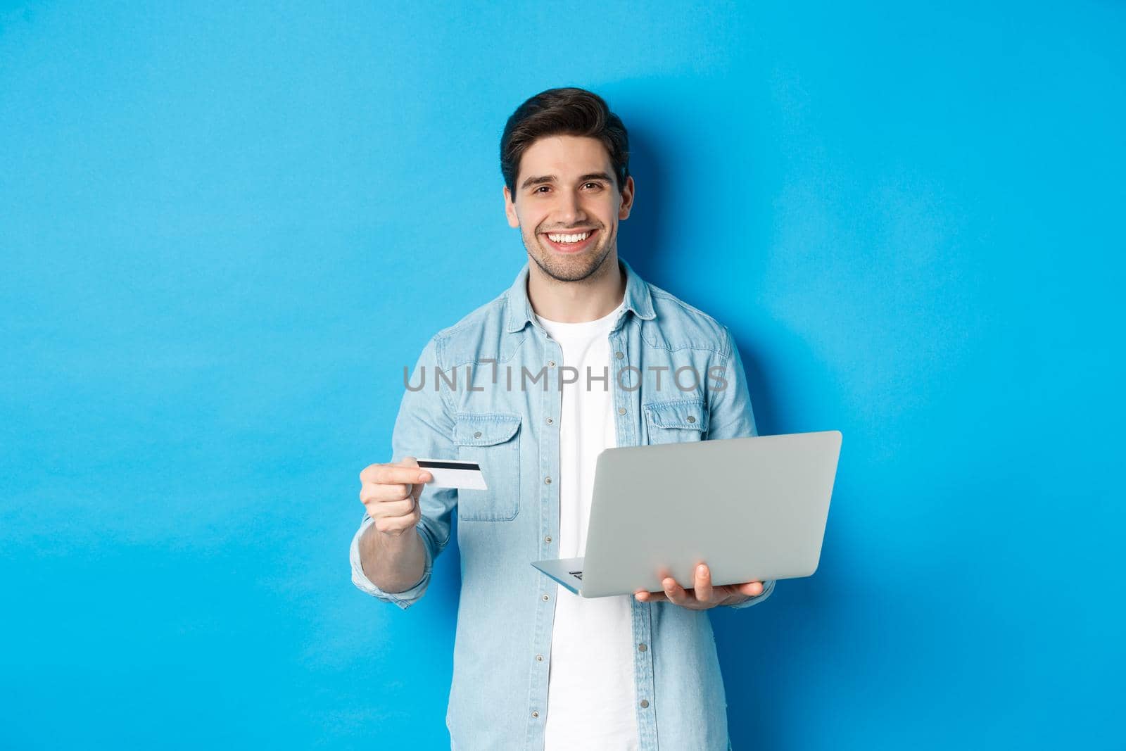 Handsome man smiling, shopping online and paying for product, holding credit card with laptop, standing over blue background.