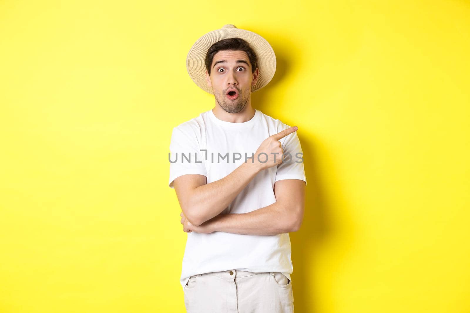 Surprised man in straw hat pointing finger right, showing promo banner, standing over yellow background by Benzoix