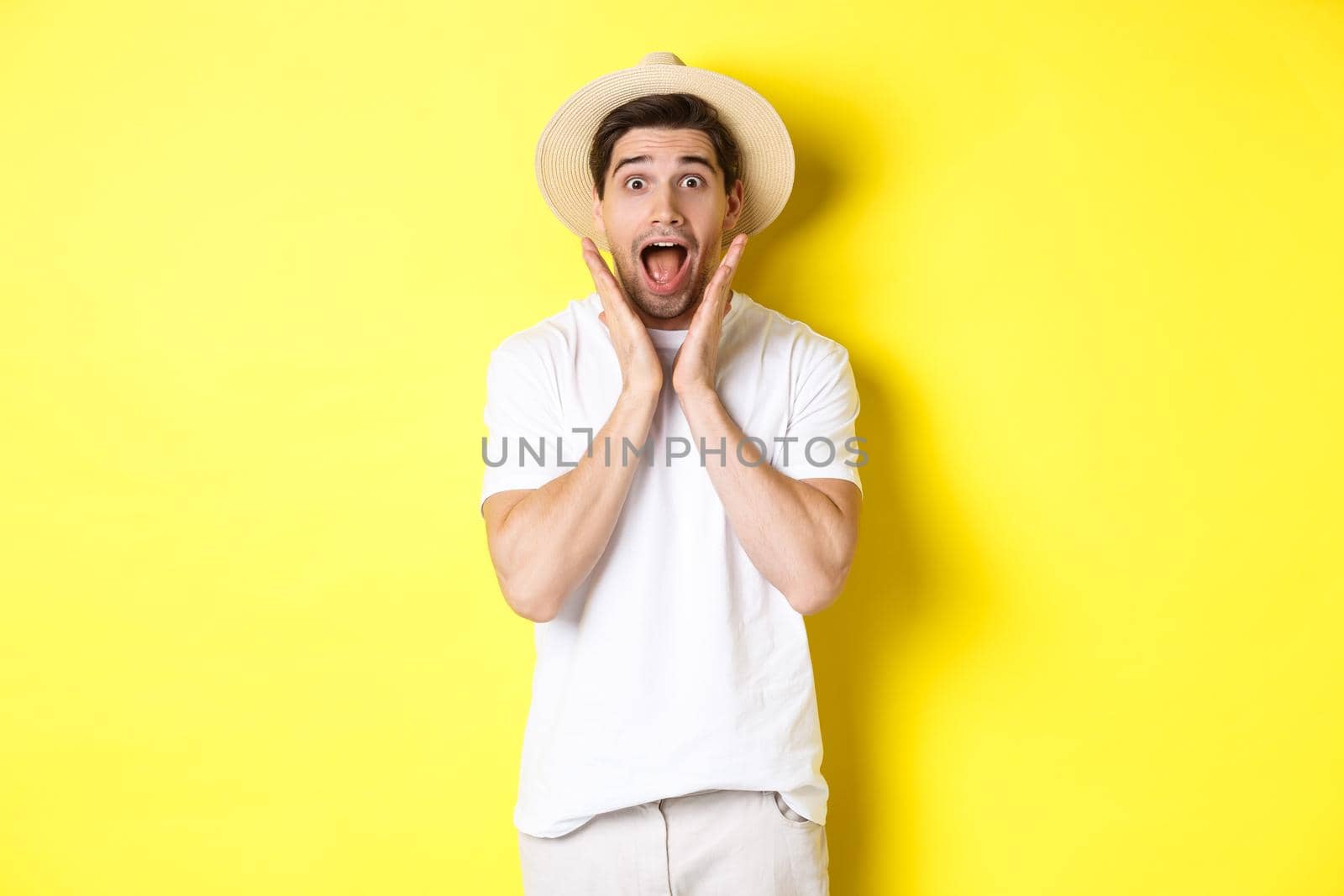 Concept of tourism and summer. Surprised male model in straw hat, looking amazed at special offer, standing against yellow background by Benzoix