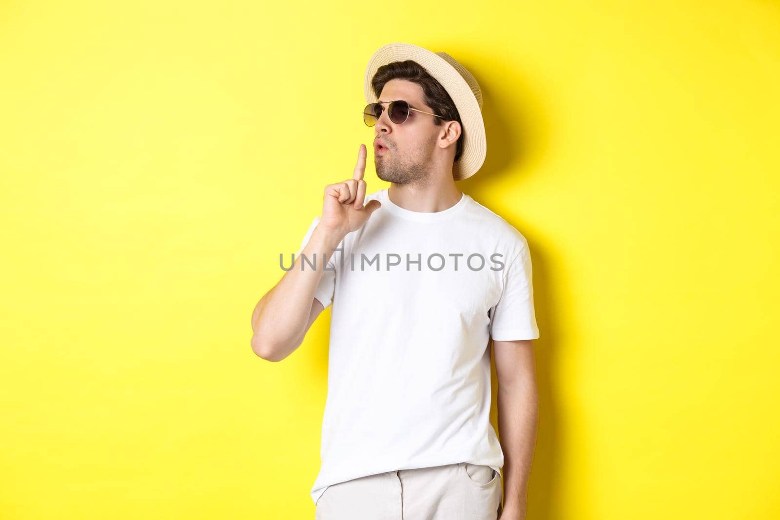 Cool young male tourist blowing at finger gun and looking confident, standing against yellow background. Vacation and lifestyle concept.