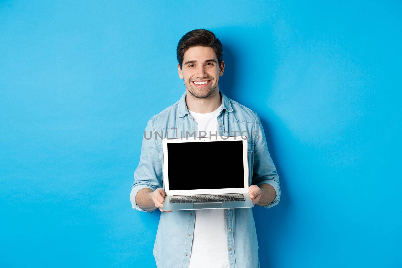 Handsome young man introduce product on laptop screen, showing computer and smiling, standing over blue background.