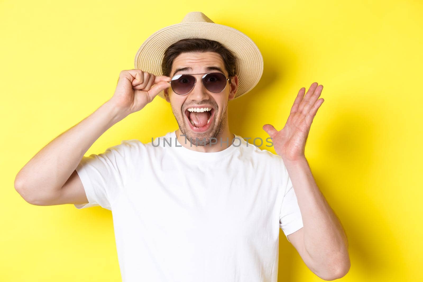 Concept of tourism and holidays. Close-up of surprised man shouting for joy, enjoying vacation, wearing sunglasses with summer hat, yellow background by Benzoix