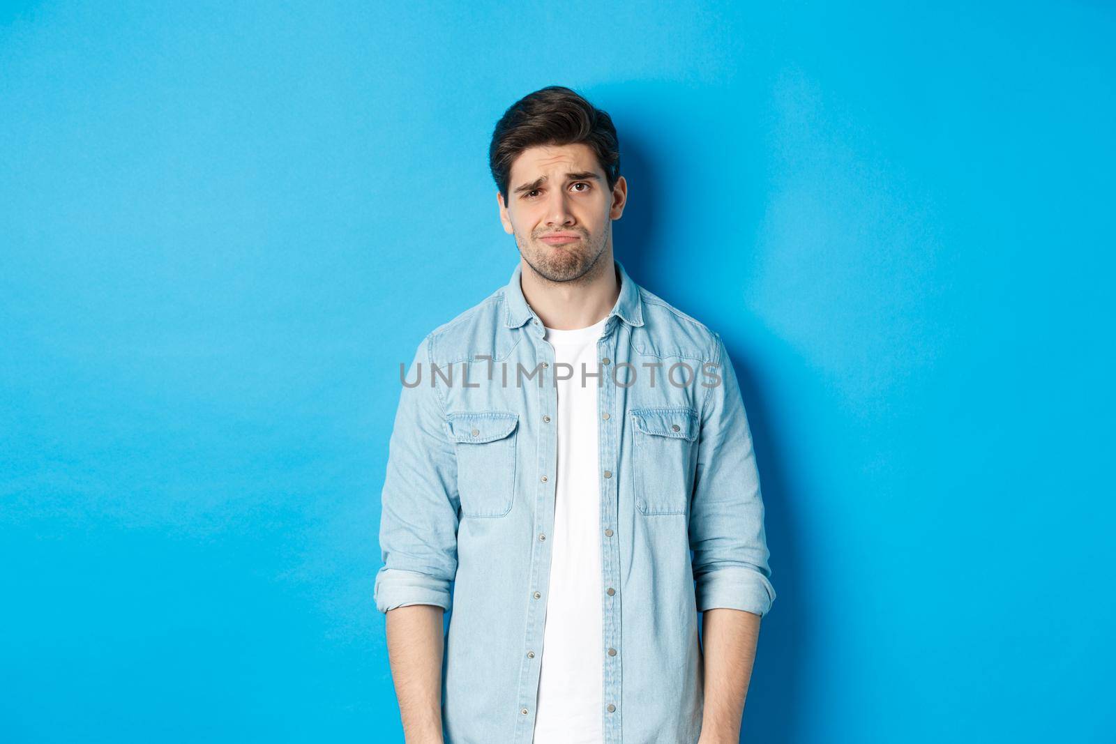 Man looking with doubt at camera, frowning and sulking, feeling uneasy about something, standing against blue background by Benzoix