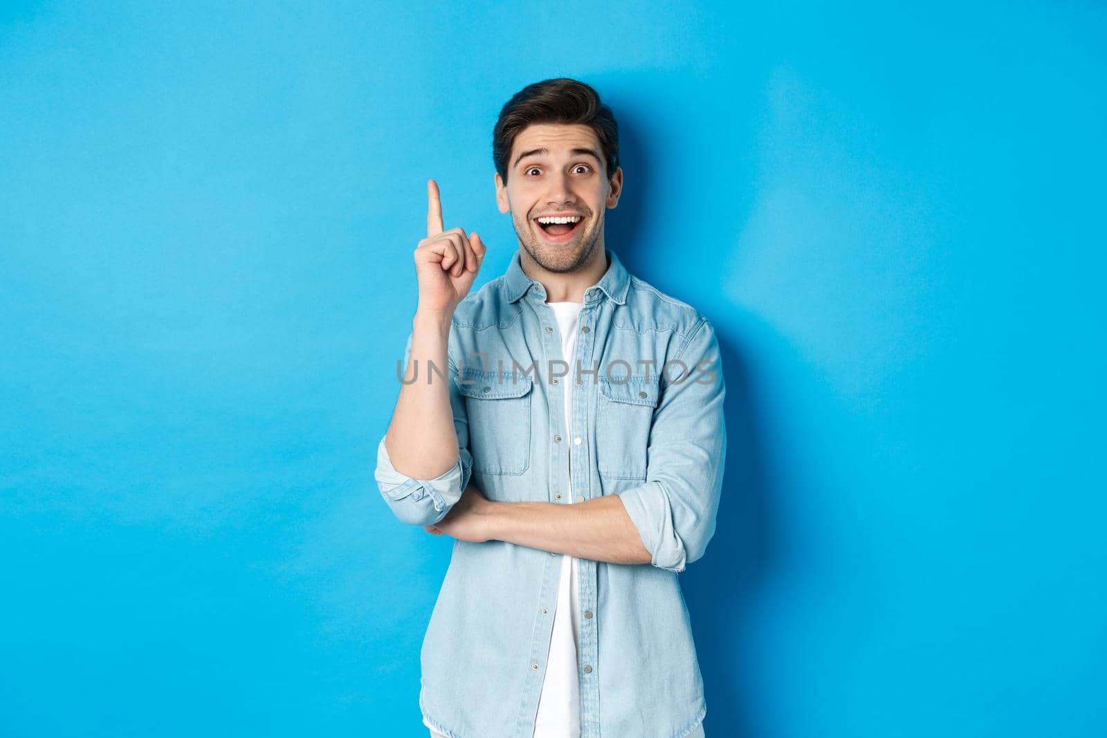 Image of handsome adult man having an idea, raising finger and smiling excited, found solution, standing against blue background.