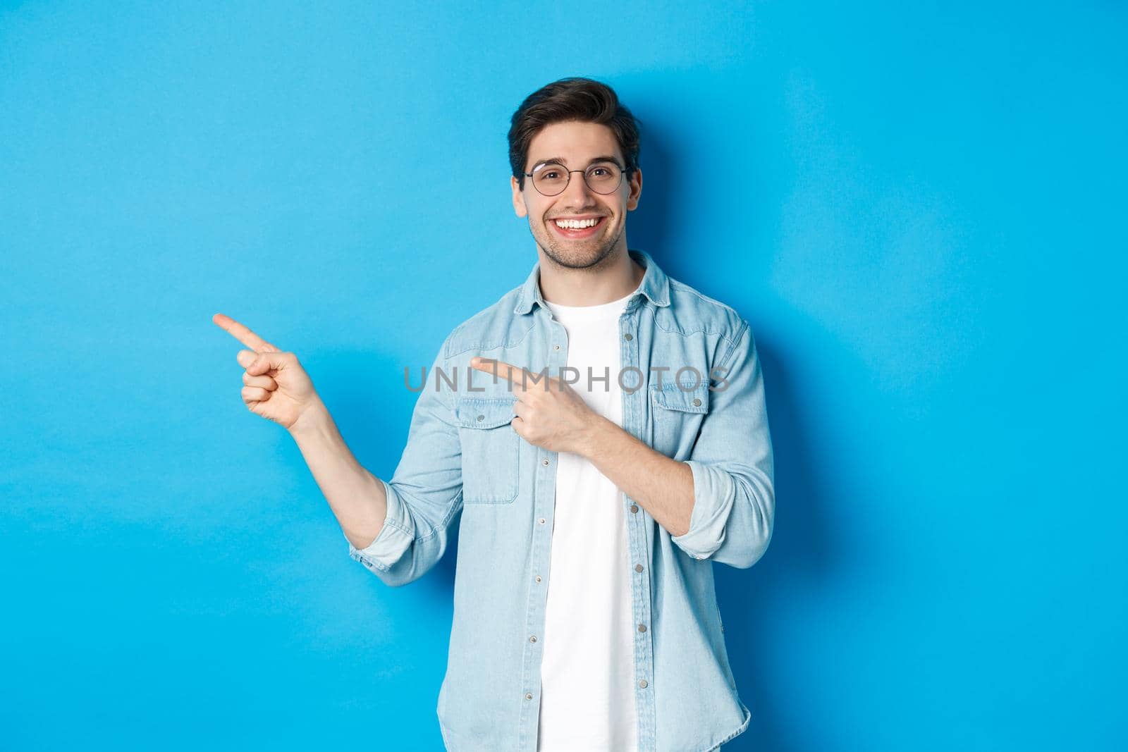 Young handsome man in glasses showing advertisement, smiling and pointing fingers left, making announcement, standing against blue background.