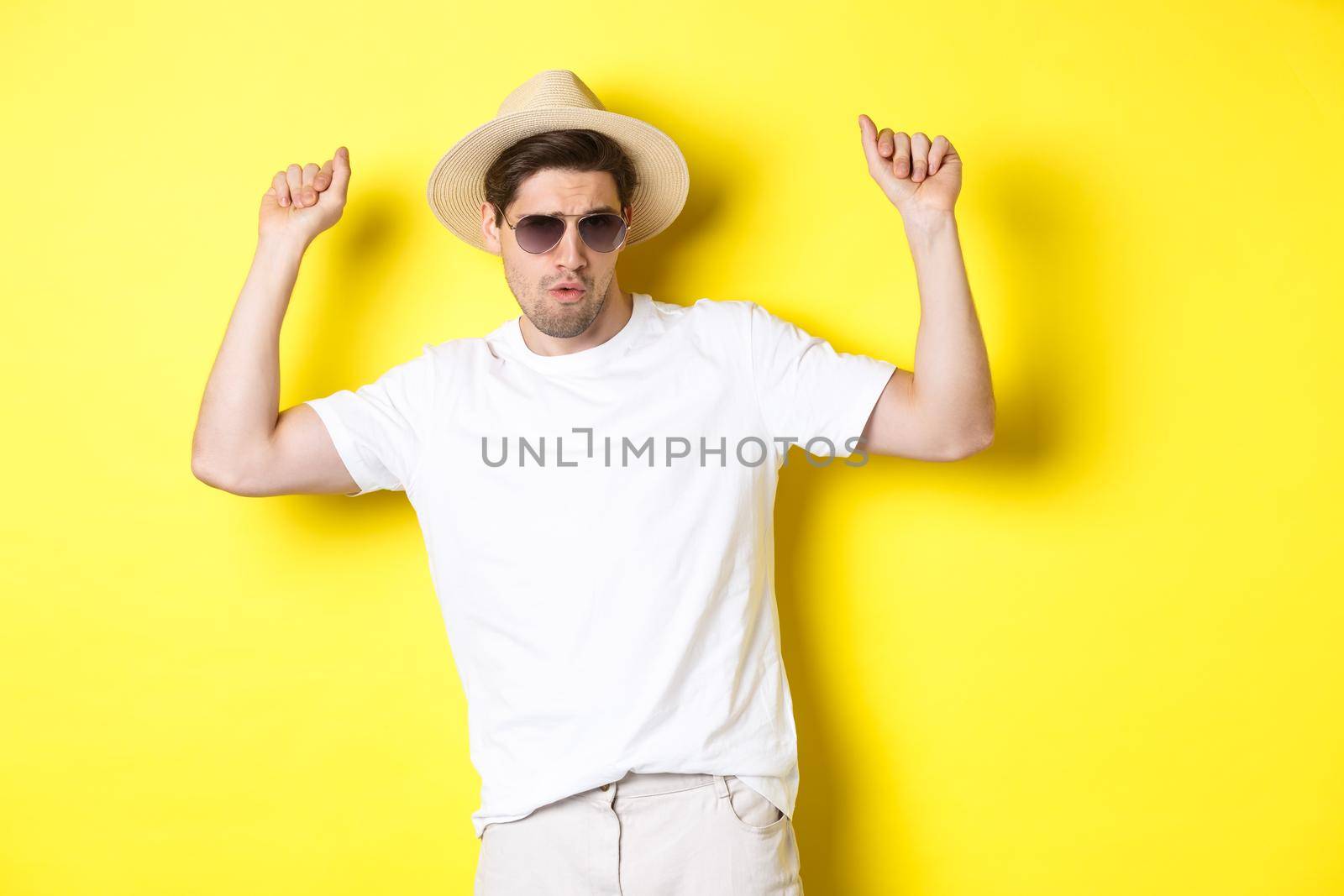 Tourism, travelling and holidays concept. Man tourist enjoying vacation, dancing in straw hat and sunglasses, posing against yellow background.