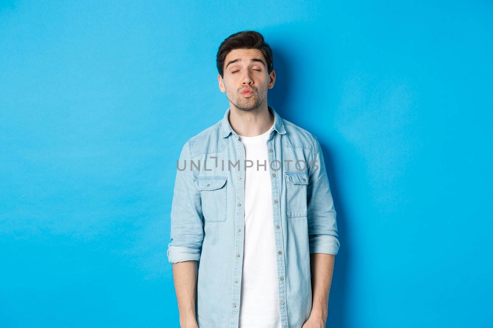 Handsome man waiting for kiss, pucker lips and close eyes while standing against blue background by Benzoix