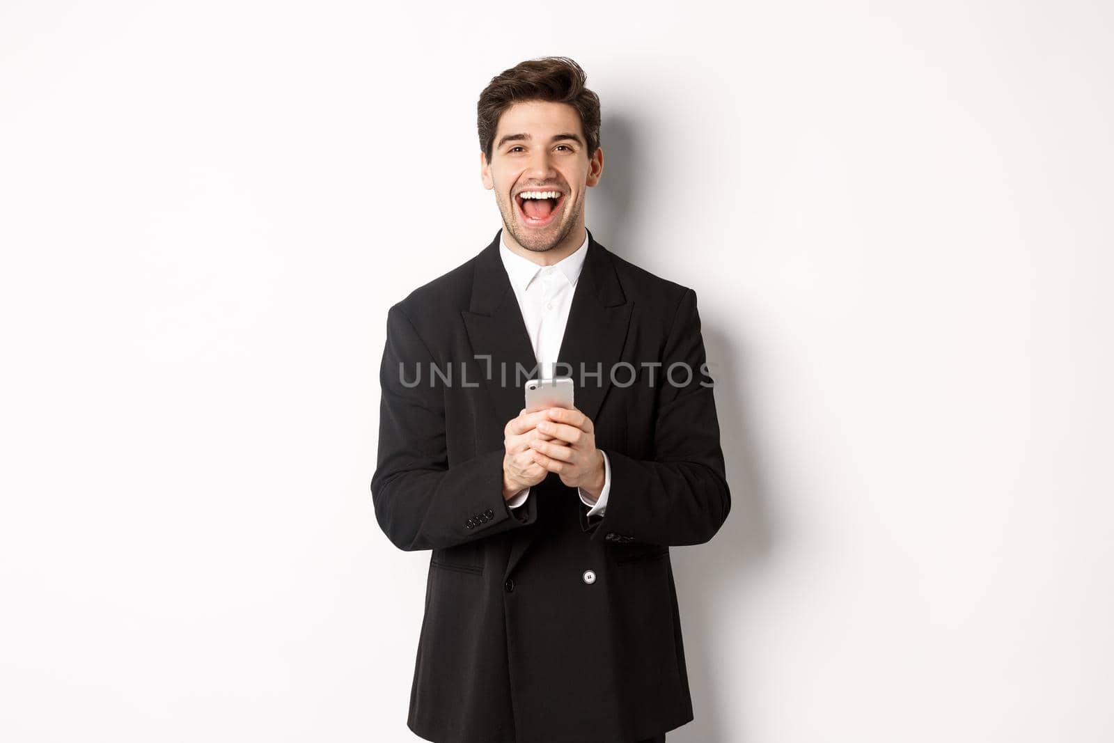 Portrait of happy good-looking man, wearing black suit, laughing from happiness and using mobile phone, standing over white background by Benzoix