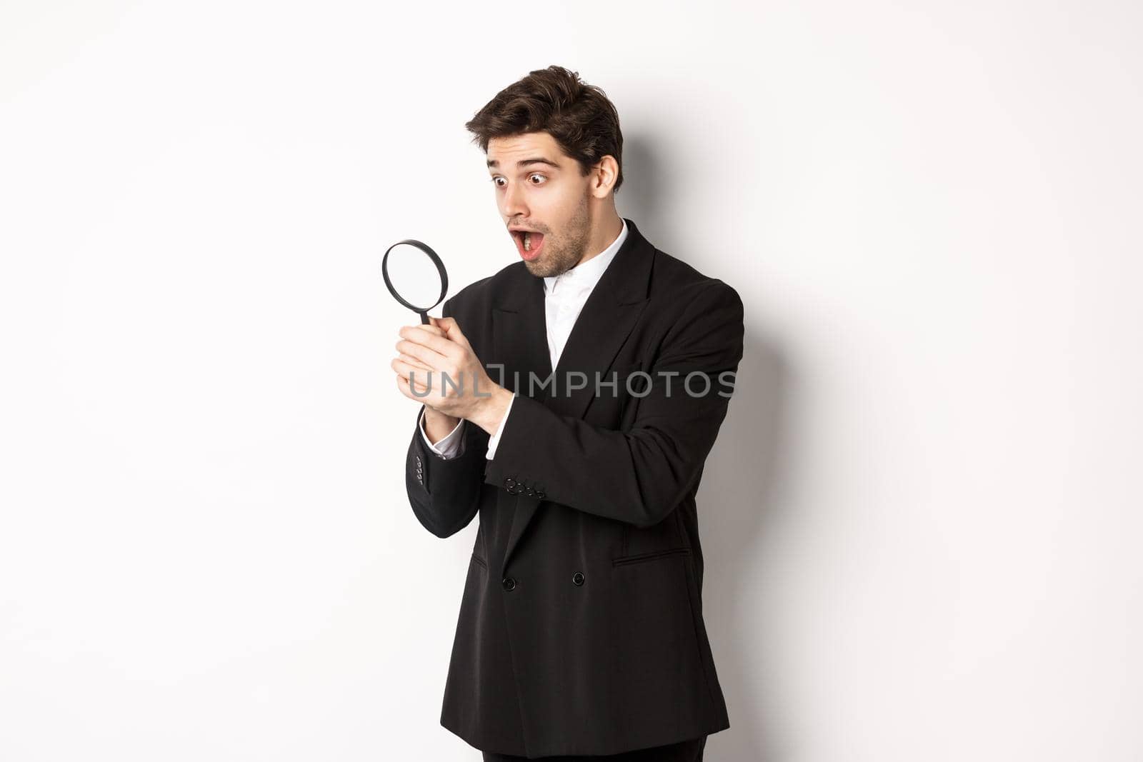 Handsome businessman in black suit, holding magnifying glass and smiling, found something, standing against white background.