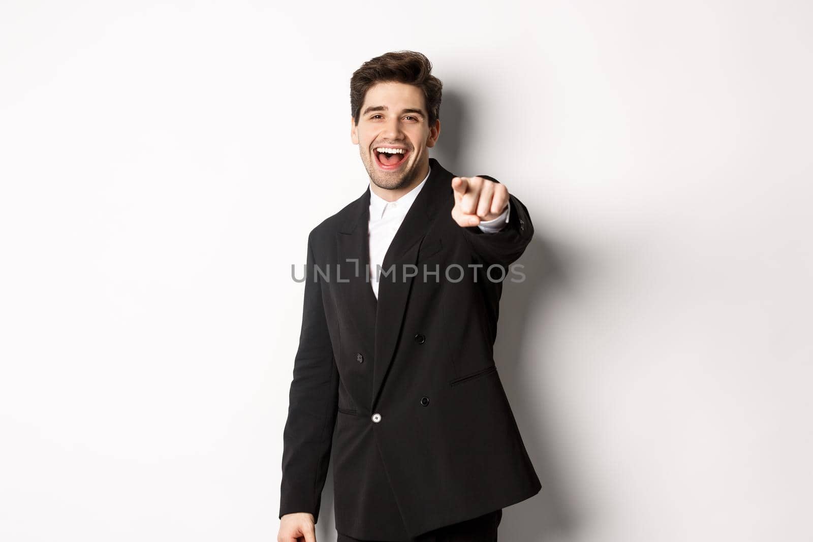 Concept of new year party, celebration and lifestyle. Portrait of handsome stylish man in black suit, smiling and pointing finger at camera, standing over white background by Benzoix