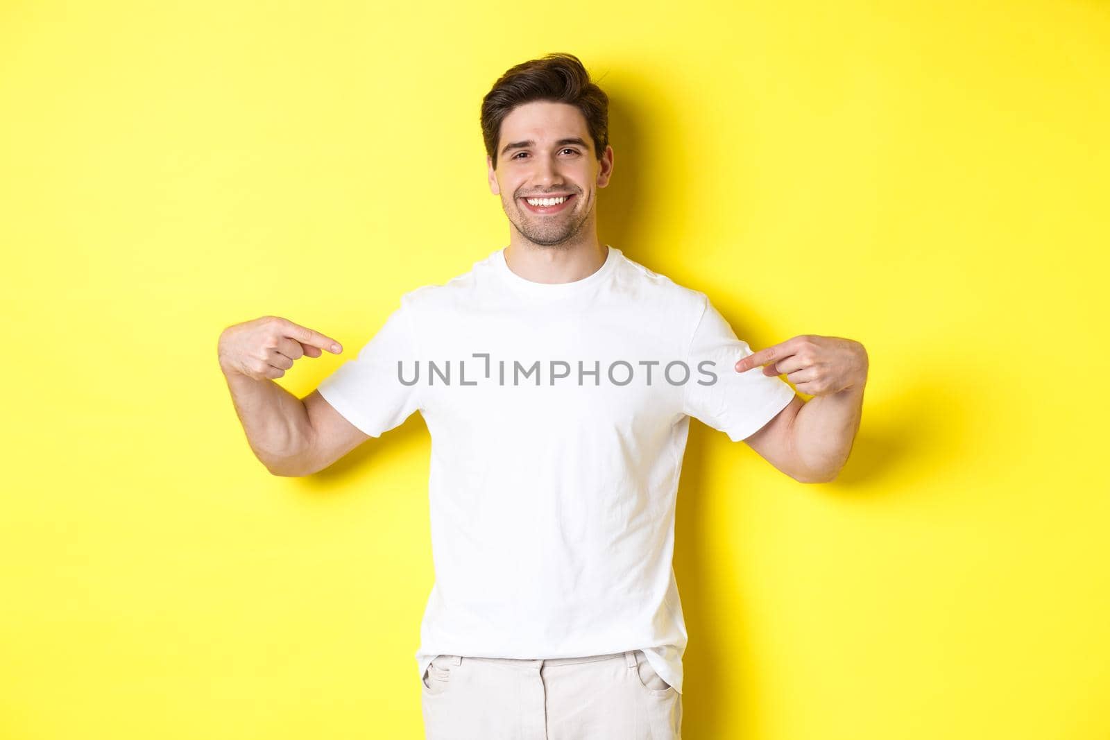 Happy attractive guy pointing fingers at your logo, showing promo on his t-shirt, standing over yellow background.