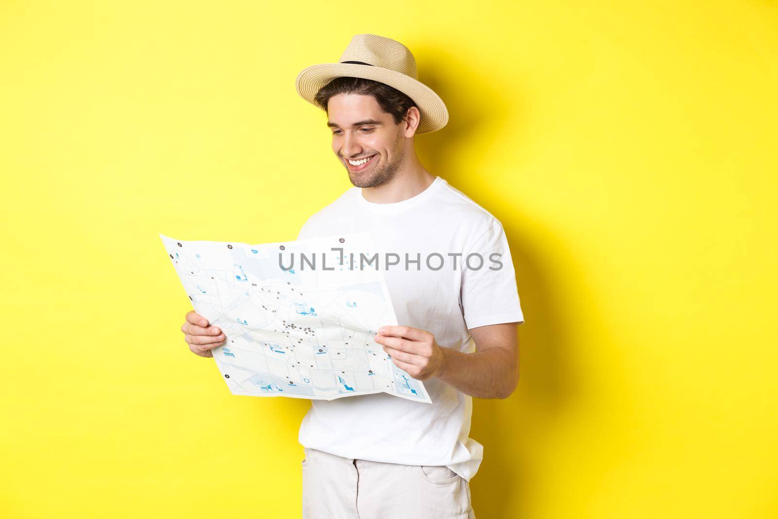 Travelling, vacation and tourism concept. Smiling happy tourist looking at map with sightseeings, standing against yellow background by Benzoix