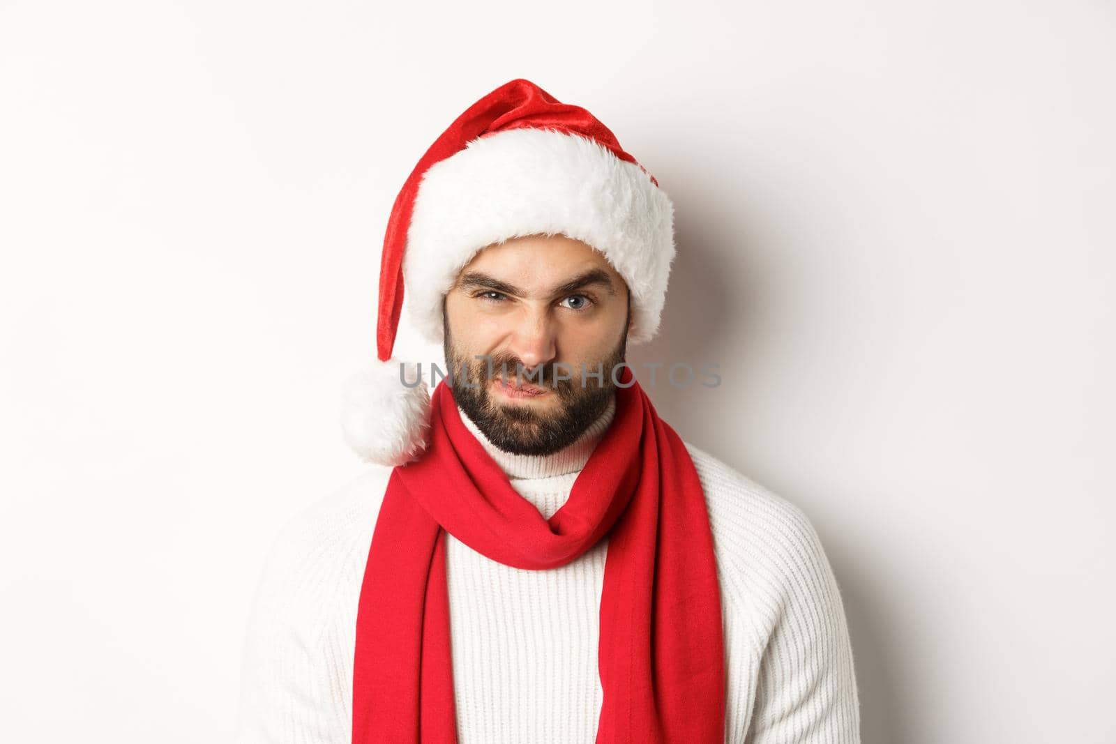 New Year party and winter holidays concept. Close-up of grumpy guy in santa hat frowning and grimacing, standing against white background.