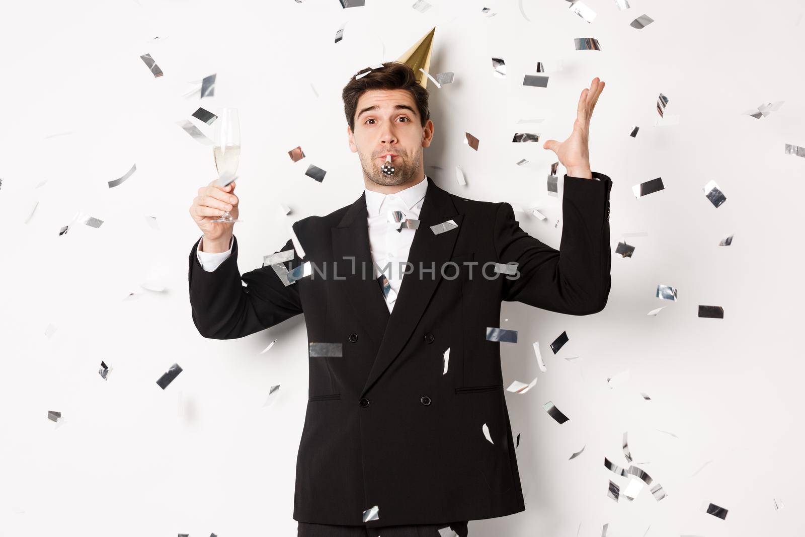 Handsome party guy in black suit having fun, celebrating new year, blowing whistle and drinking champagne while confetti falling, wishing happy holidays, standing against white background by Benzoix