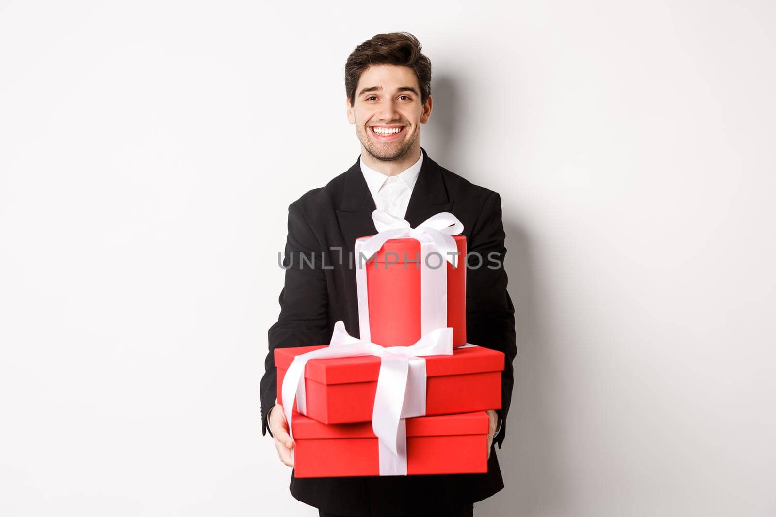 Portrait of handsome bearded man in trendy suit, holding gifts for new year and smiling, prepared presents, standing over white background.