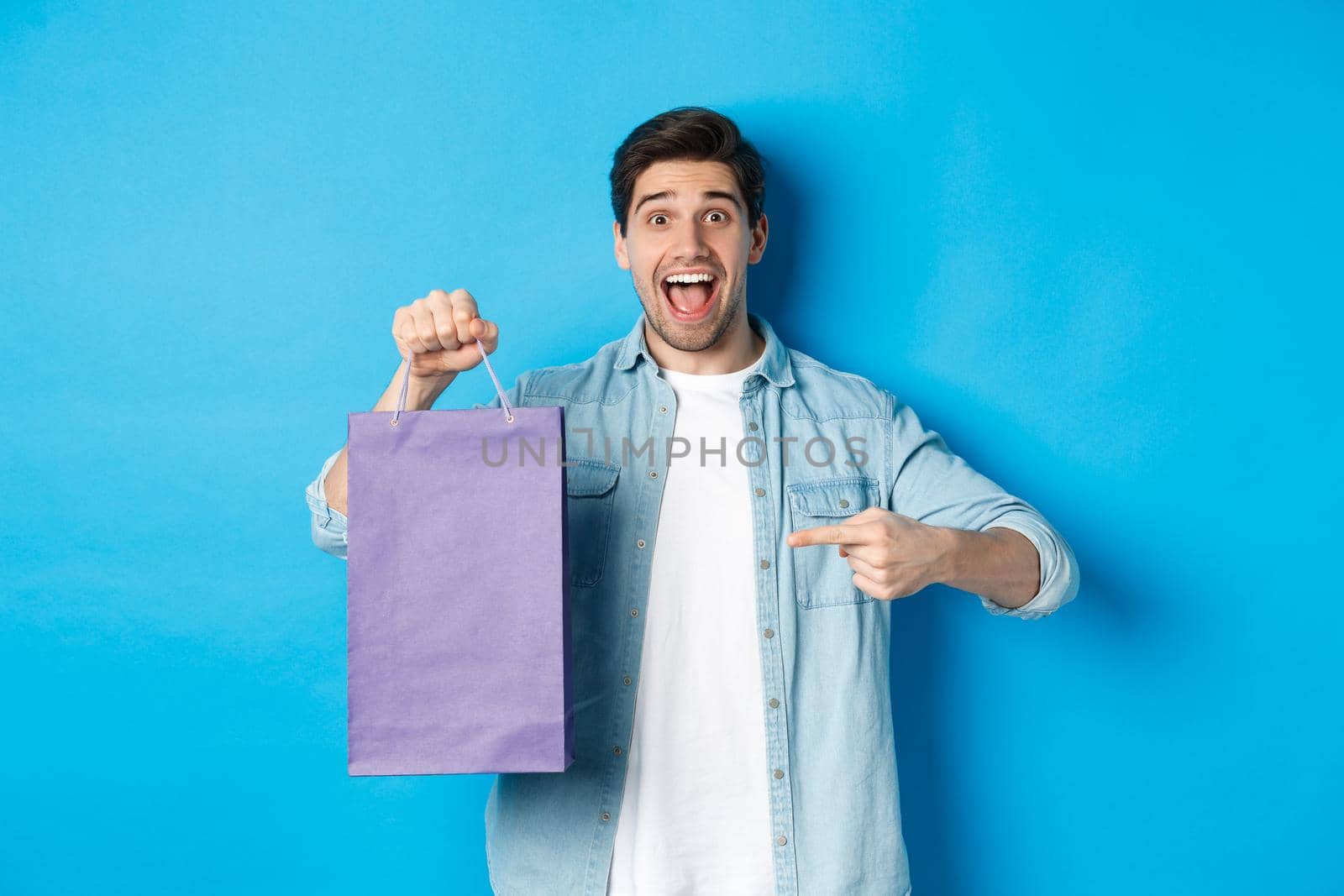 Concept of shopping, holidays and lifestyle. Excited guy pointing finger at paper bag and looking amazed, recommending store, announcing discounts, blue background.