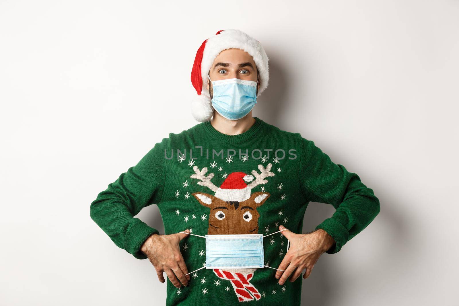Concept of covid-19 and Christmas holidays. Funny guy put on face mask on his sweater deer, standing over white background.