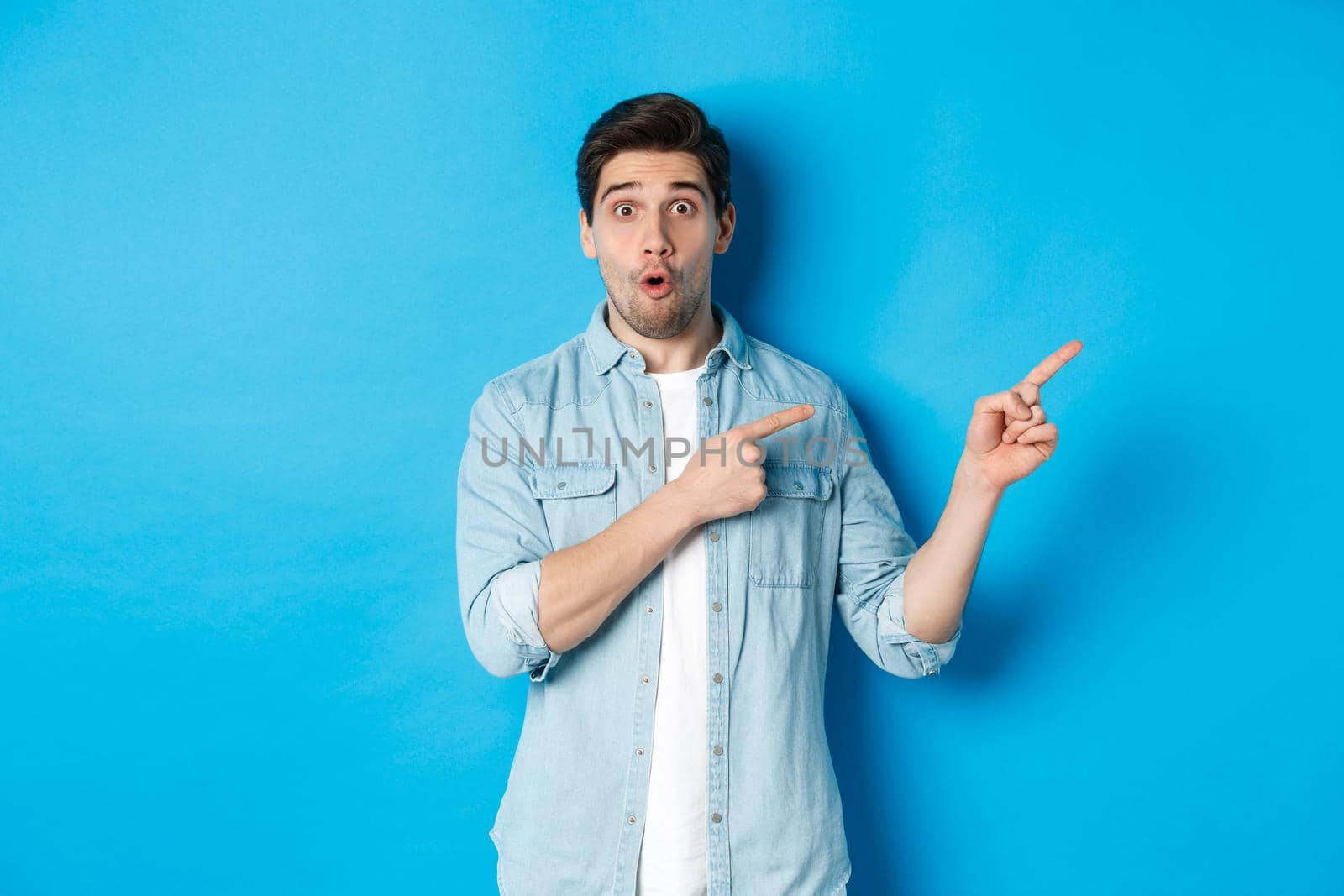 Portrait of surprised bearded guy in casual outfit pointing fingers right, showing amazing promo offer, standing over blue background.