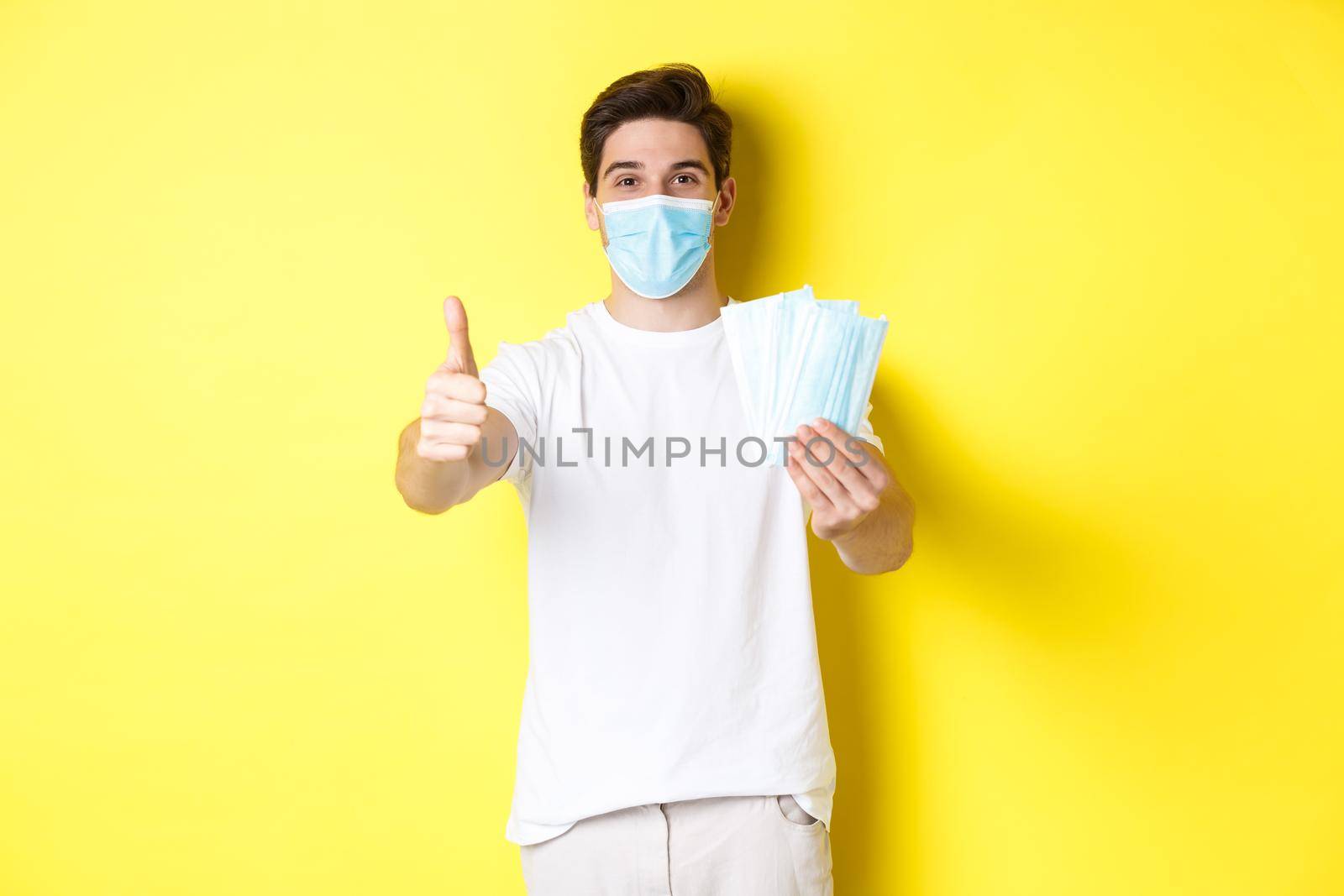 Concept of covid-19, quarantine and preventive measures. Satisfied man showing thumb up and giving medical masks, standing over yellow background.