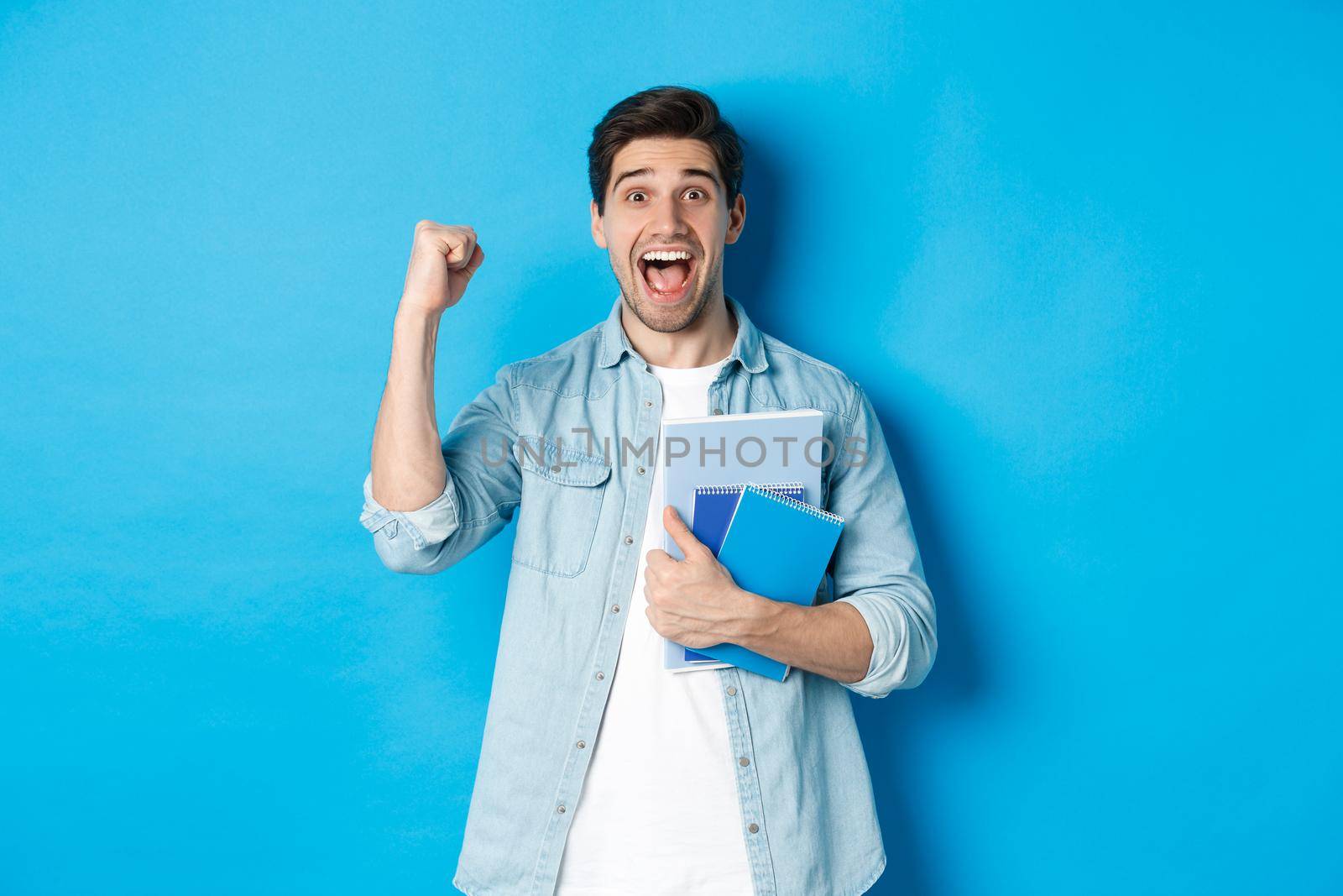 Cheerful guy holding notebooks and celebrating, making fist pump and shouting yes with excitement, standing over blue background by Benzoix