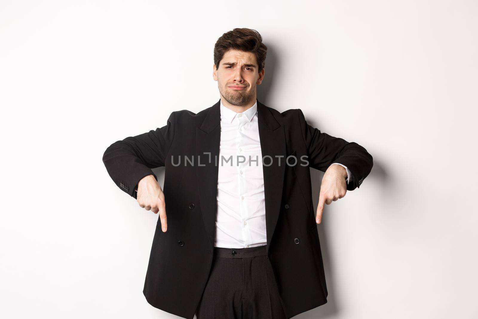 Portrait of skeptical and unamused bearded man, pointing fingers down with disappointed face, standing in suit and complaining.