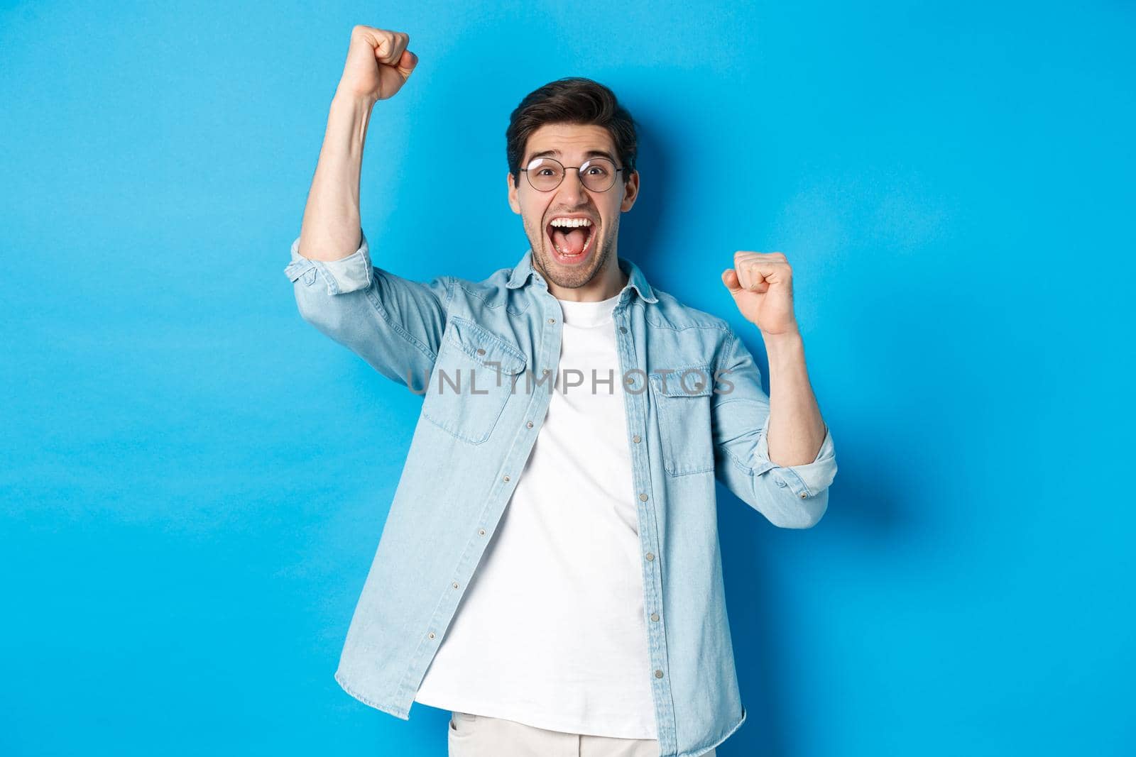 Excited handsome man triumphing, raising hands up and shouting for joy, celebrating victory, standing against blue background by Benzoix