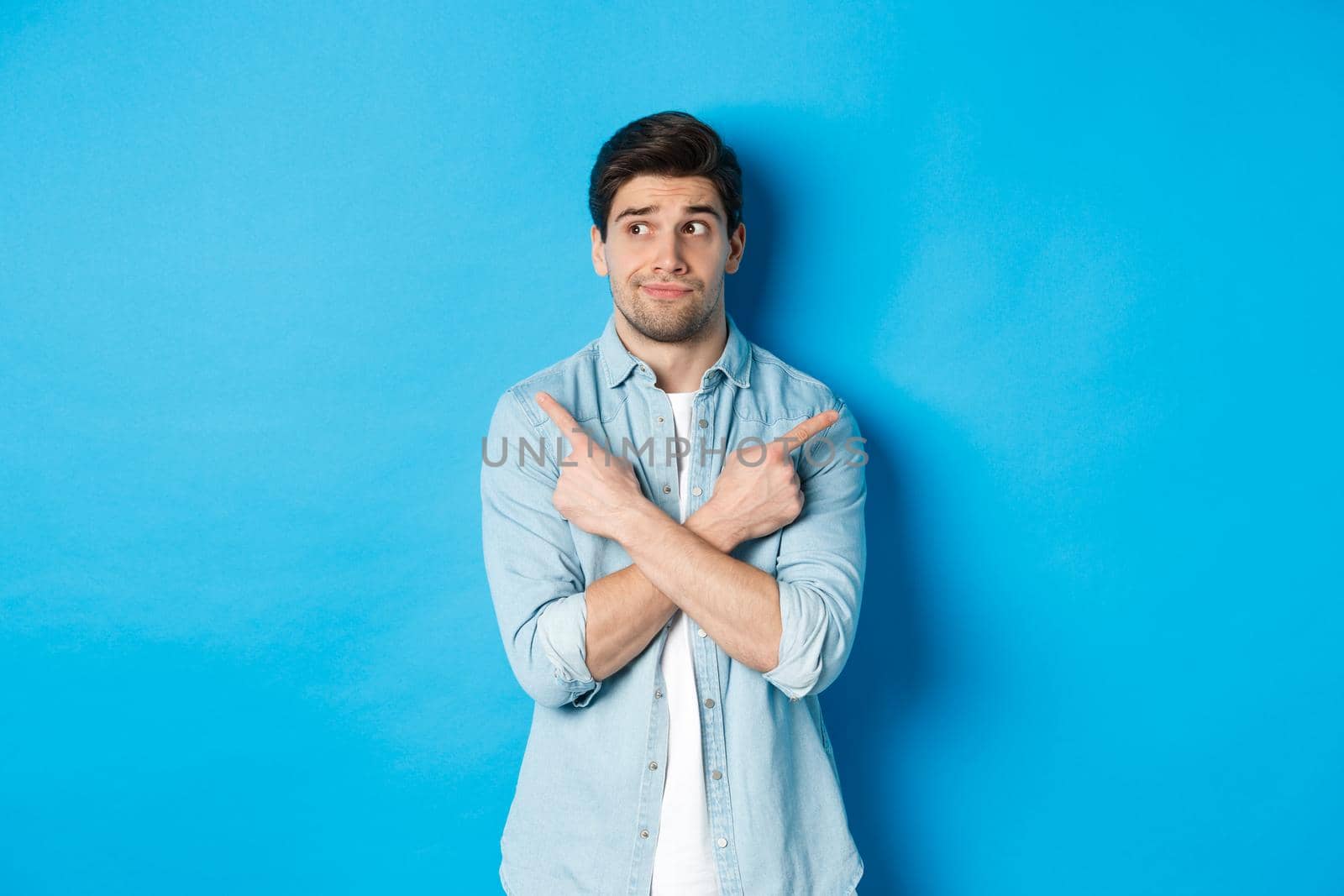 Portrait of indecisive adult man pointing fingers sideways but looking left, making choice between two products, standing against blue background by Benzoix