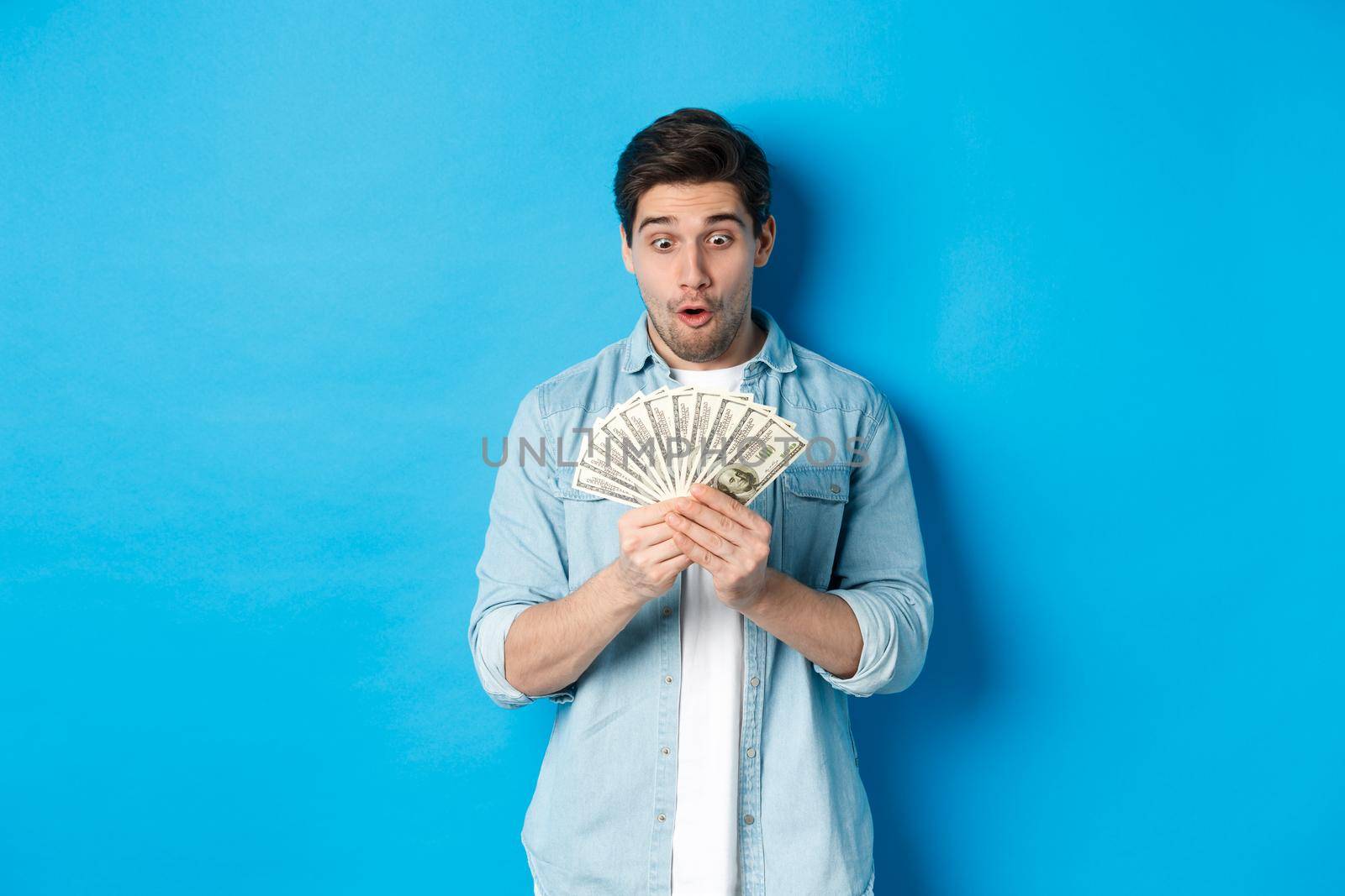 Excited successful man counting money, looking satisfied at cash and smiling, standing over blue background by Benzoix