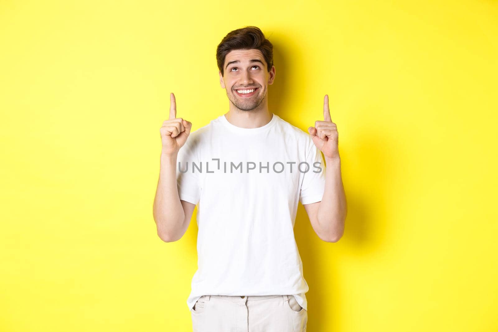 Excited attractive man in white t-shirt, pointing fingers up, looking at advertisement with happy smile, standing over yellow background by Benzoix