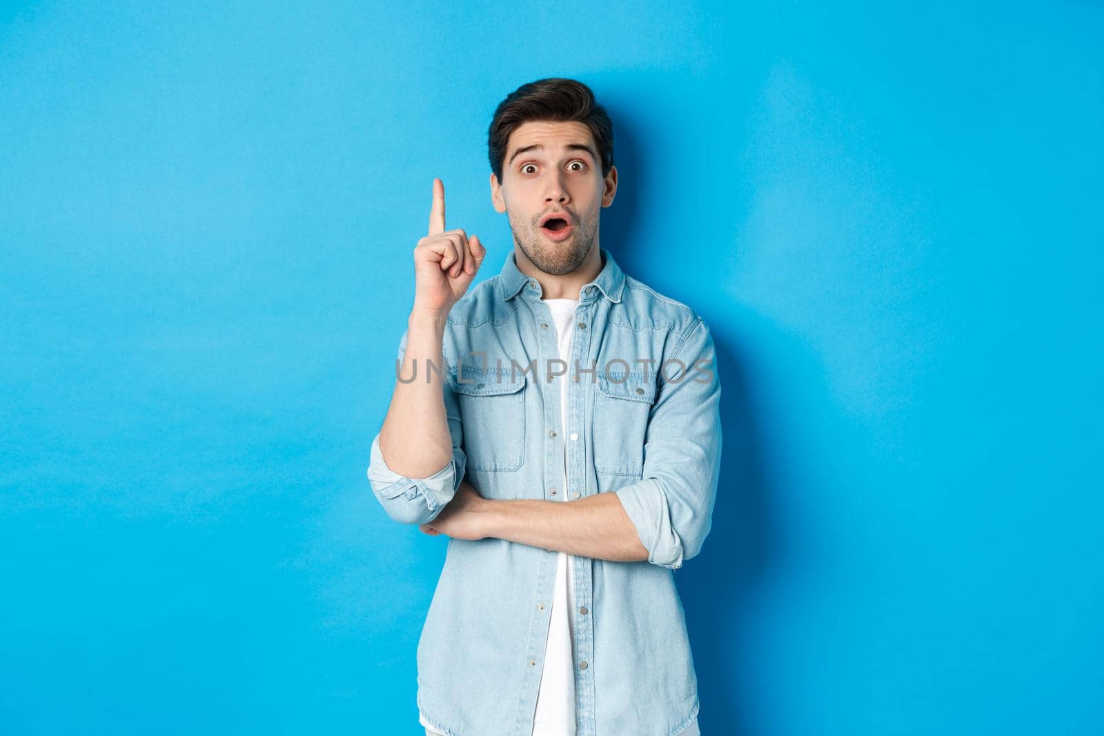 Image of handsome adult man having an idea, raising finger and smiling excited, found solution, standing against blue background by Benzoix