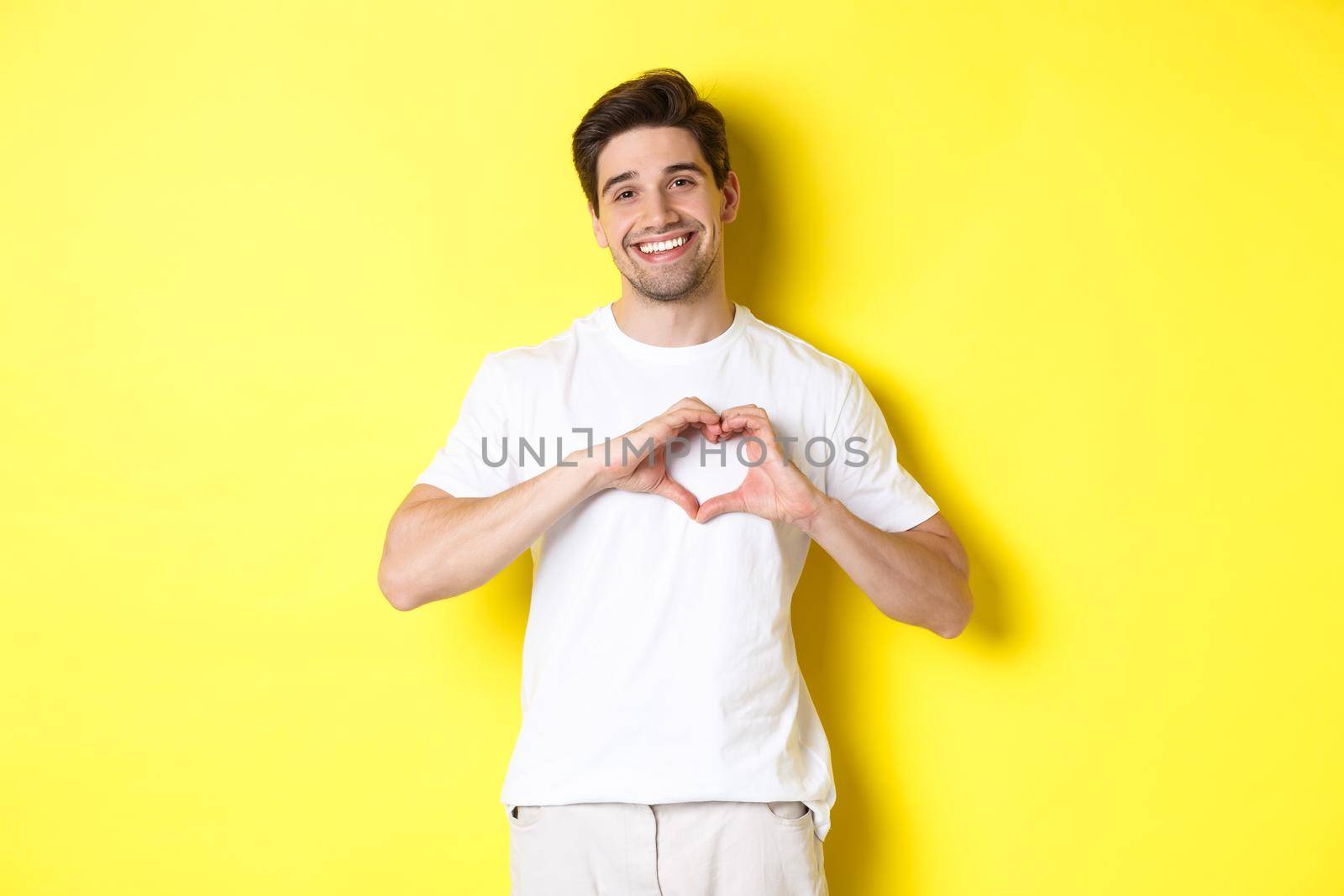 Happy romantic man showing heart sign, smiling and express love, standing over yellow background by Benzoix
