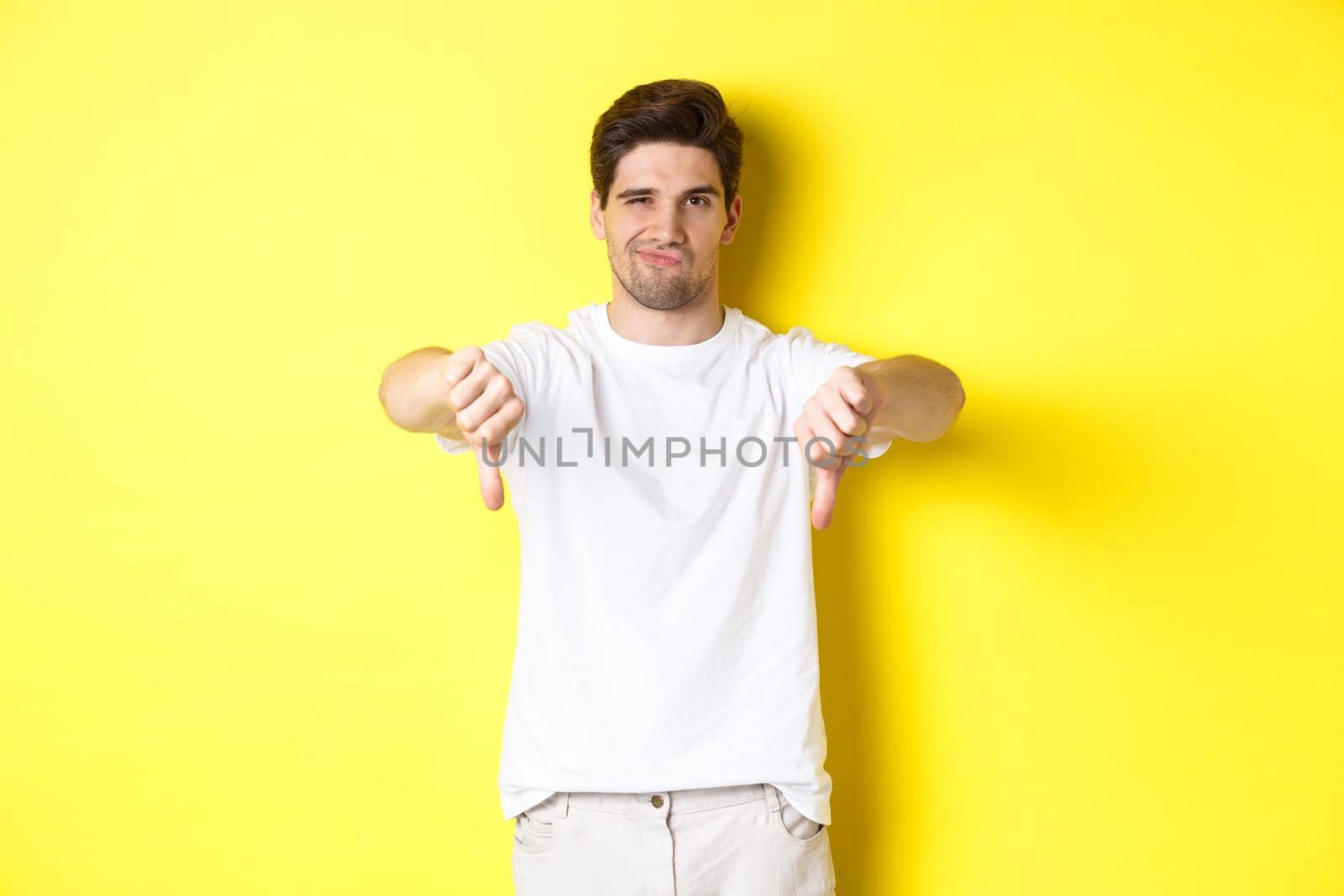 Skeptical guy showing thumbs down and smirking, dislike and disagree, standing over yellow background. Copy space