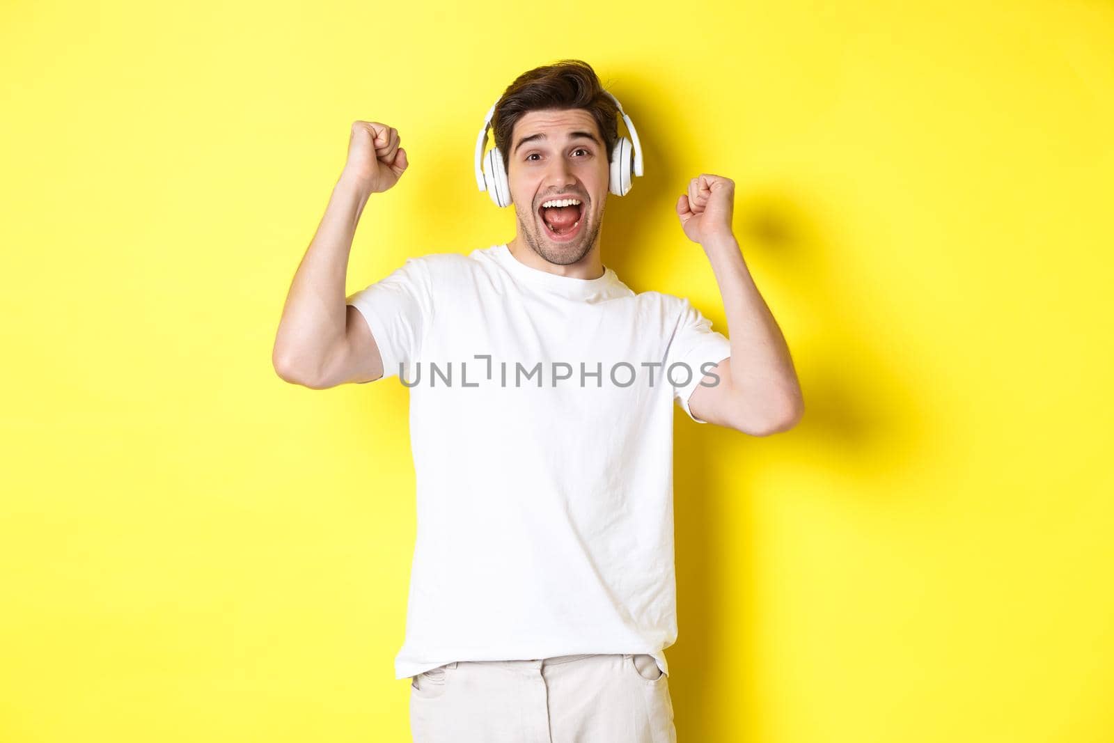 Excited handsome man dancing and singing along, listening music in headphones, standing over yellow background by Benzoix