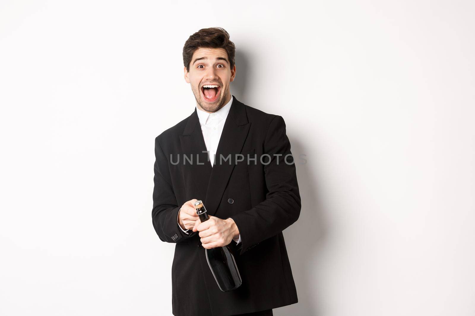 Image of attractive man in black suit having a party, celebrating new year and opening bottle of champagne, standing happy against white background.